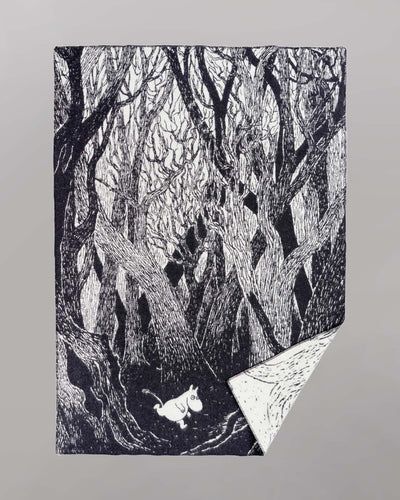 A black and white Klippan Moomin on the Run Blanket featuring an illustration of dense, intertwined trees in a forest with a small white creature walking along the forest floor. This eco-friendly children's blanket is crafted from organic lambswool, ensuring both comfort and sustainability.