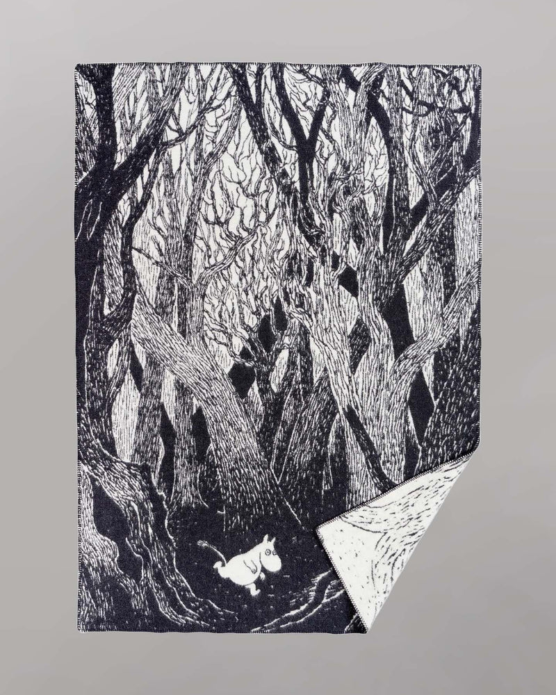 A black and white Klippan Moomin on the Run Blanket featuring an illustration of dense, intertwined trees in a forest with a small white creature walking along the forest floor. This eco-friendly children's blanket is crafted from organic lambswool, ensuring both comfort and sustainability.