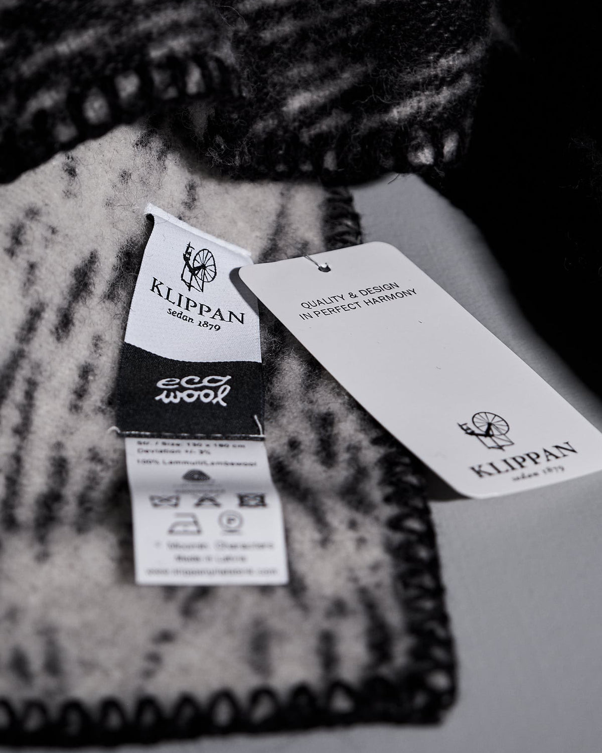 Close-up of a black and white woven fabric with three tags attached. One tag displays the brand "Klippan," the second tag reads "eco wool," and the third tag highlights Swedish craftsmanship, quality, and design. This Moomin on the Run Blanket by Klippan exemplifies refined eco-friendly children's products.