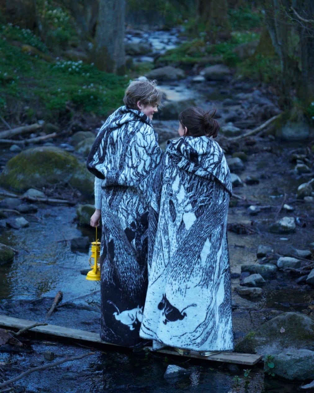 Two people wrapped in eco-friendly Moomin on the Run Blankets by Klippan stand on a small wooden plank bridge over a rocky stream in the forest, one holding a yellow lantern.