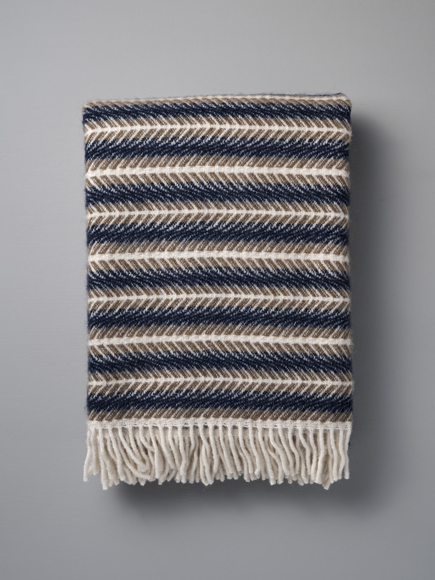 The Klippan Oscar Wool Blanket – Blue Sand features a navy, brown, and cream chevron pattern with cream fringe, beautifully folded against a gray background—a true luxury throw.