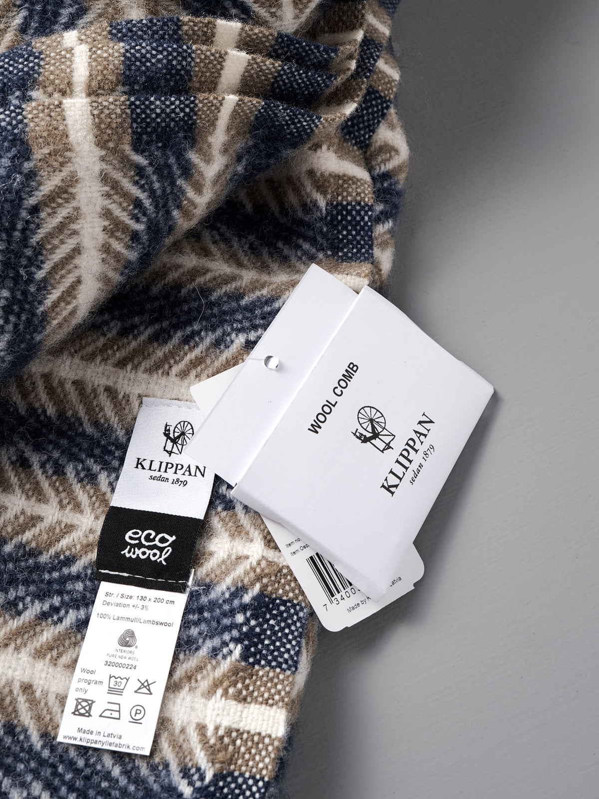 A close-up of the Klippan Oscar Wool Blanket – Blue Sand, highlighting its blue and beige patterns along with attached Klippan brand tags and a wool comb label.