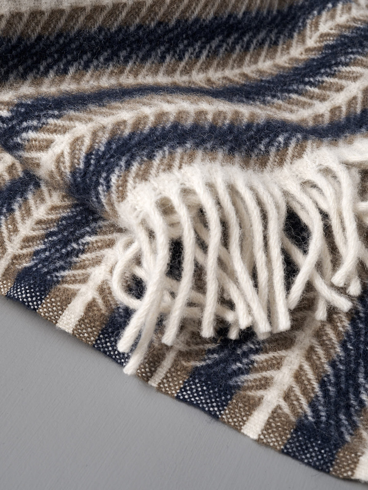 Close-up of the Klippan Oscar Wool Blanket – Blue Sand, a Swedish wool throw with blue and sand stripes and fringed edges, displayed on a gray surface.