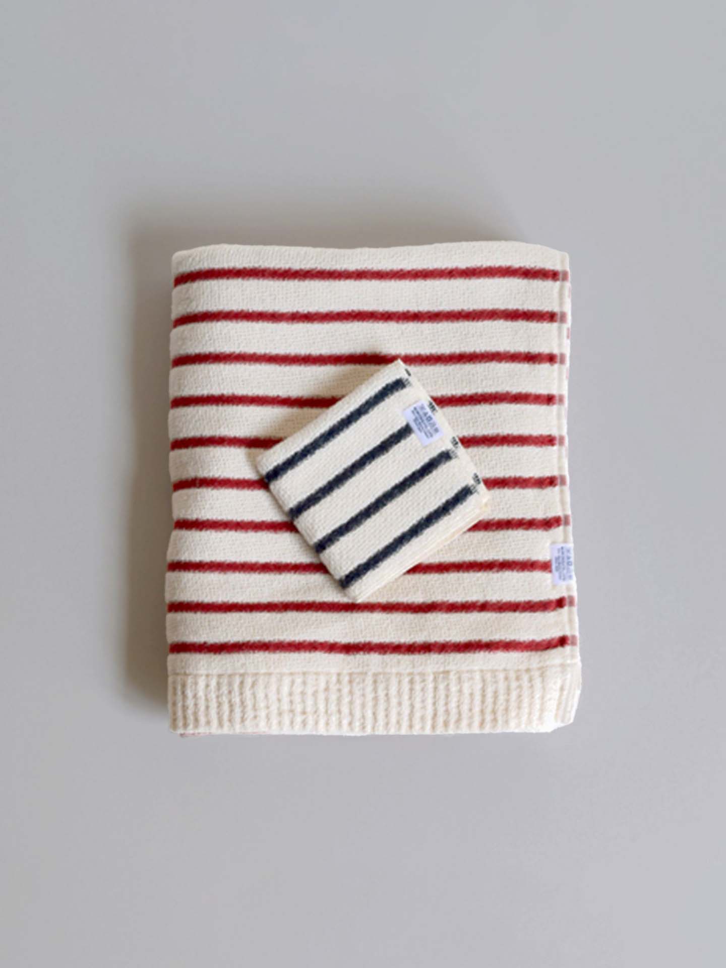 A folded beige towel featuring red stripes, crafted from 100% cotton, rests next to a smaller beige washcloth with dark blue stripes on a light gray surface. These Kids Stripe Towels by Kontex are perfect for gentle use on children's and babies' skin and are meticulously made in Imabari, Japan.