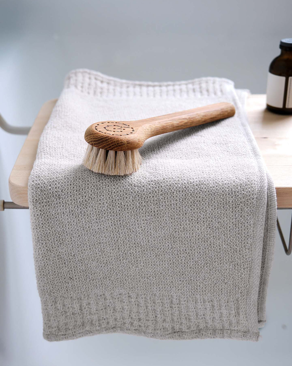 A Re.Lana towel from Kontex, with its soft texture, is gently folded atop a wooden bench alongside a wooden bath brush. In the background, a small brown bottle enhances the tranquil ambiance.