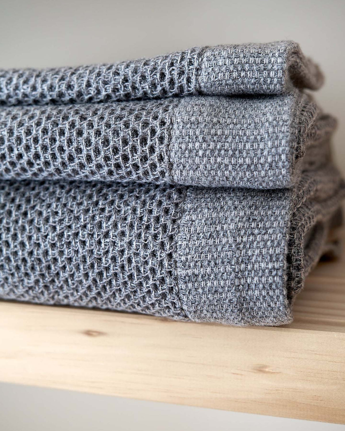 Three folded gray knit blankets stacked on a wooden shelf, accompanied by a Kontex Vintage Waffle Towel recognized for its luxurious softness.