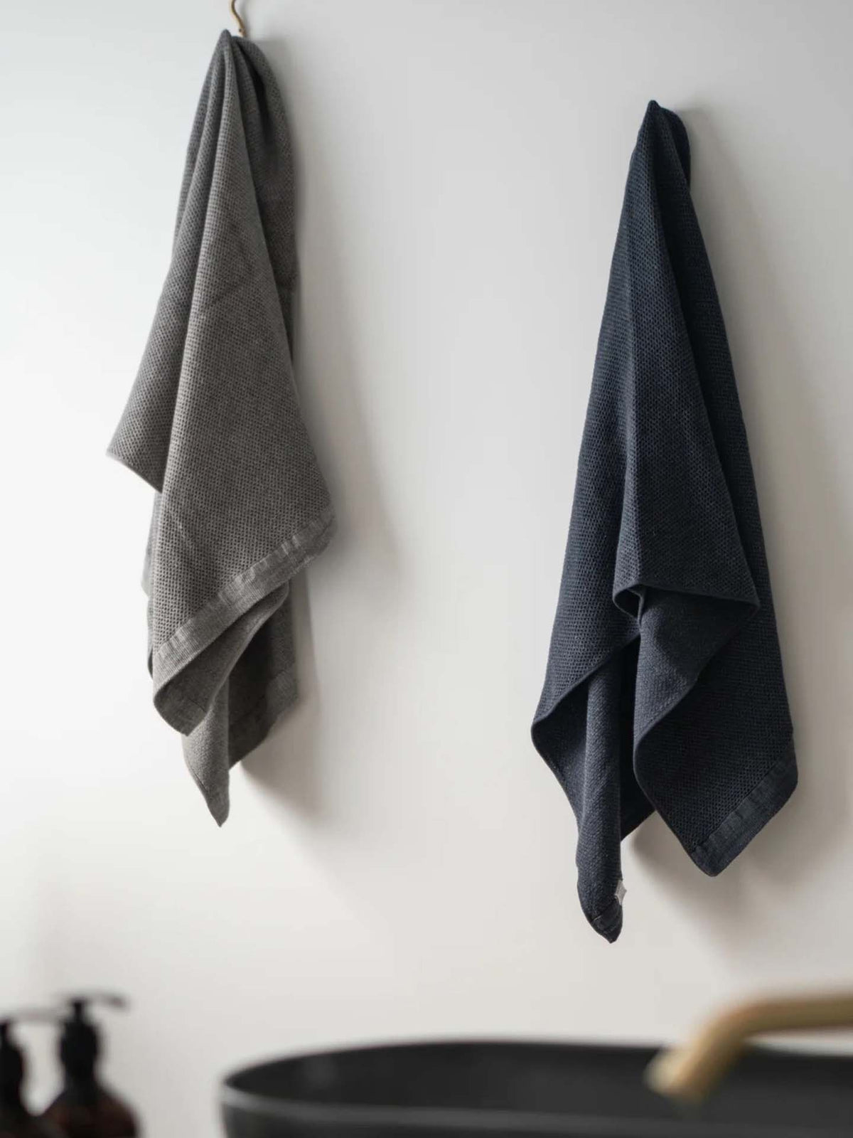 Two Vintage Waffle Towels by Kontex, one in a gray Imabari design and the other in a dark blue linen cotton blend, hang on hooks against a white wall near a sink.