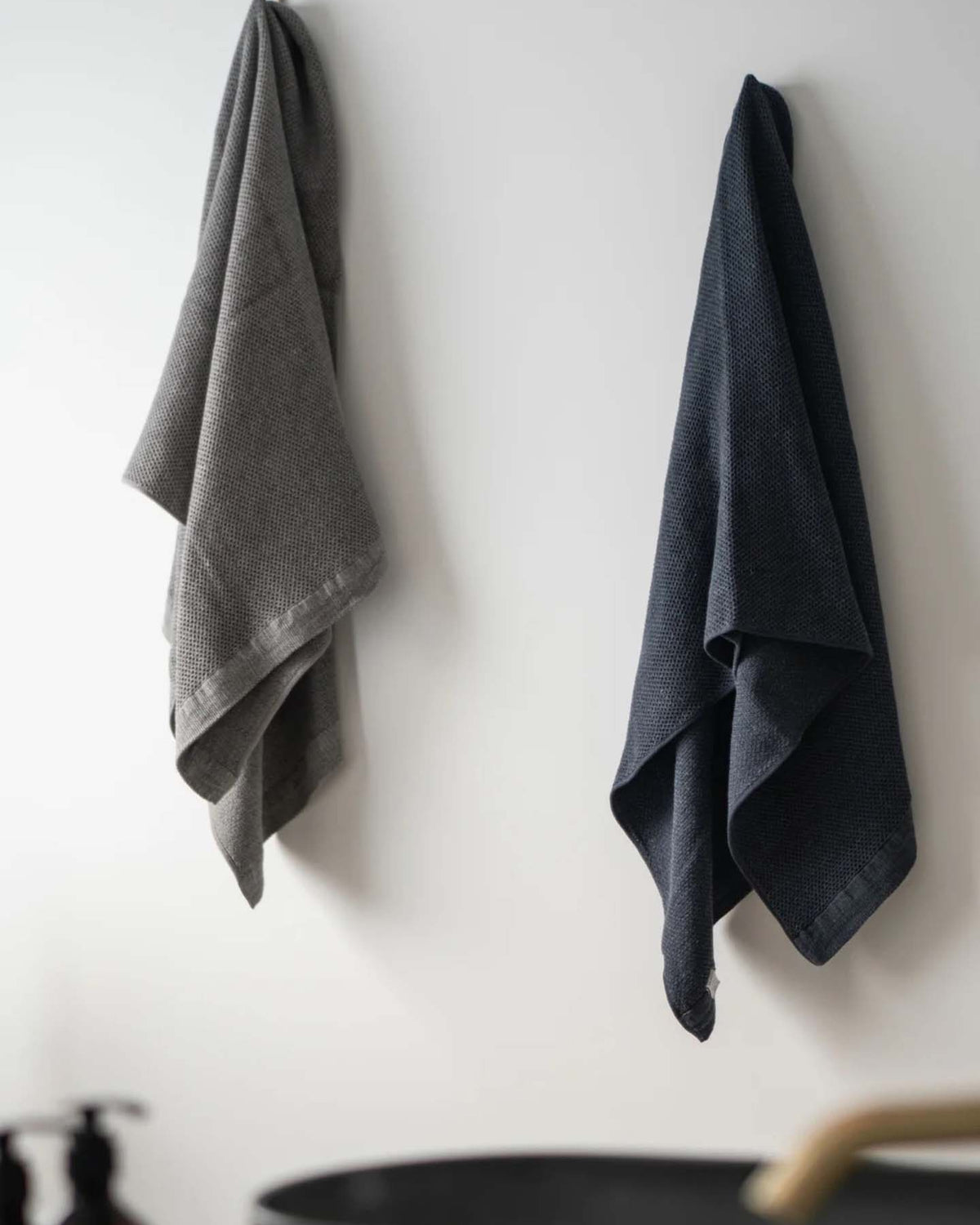 Two Vintage Waffle Towels by Kontex, one in a gray Imabari design and the other in a dark blue linen cotton blend, hang on hooks against a white wall near a sink.