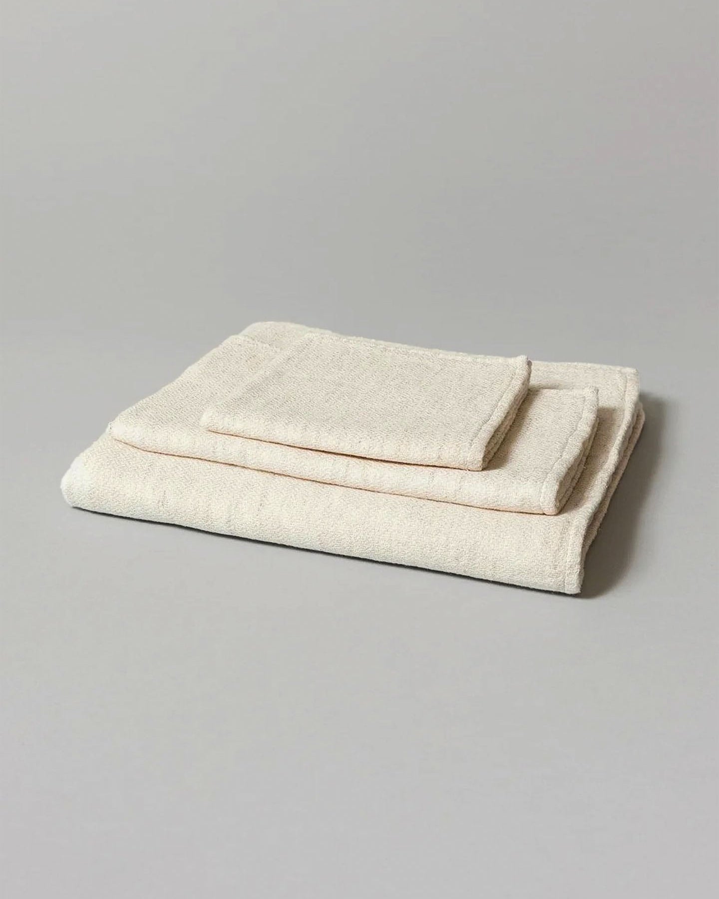 Two neatly folded Claire towels from Kontex, crafted from organic cotton and featuring one large bath towel with a smaller fast-drying hand towel on top, rest on a plain gray surface.