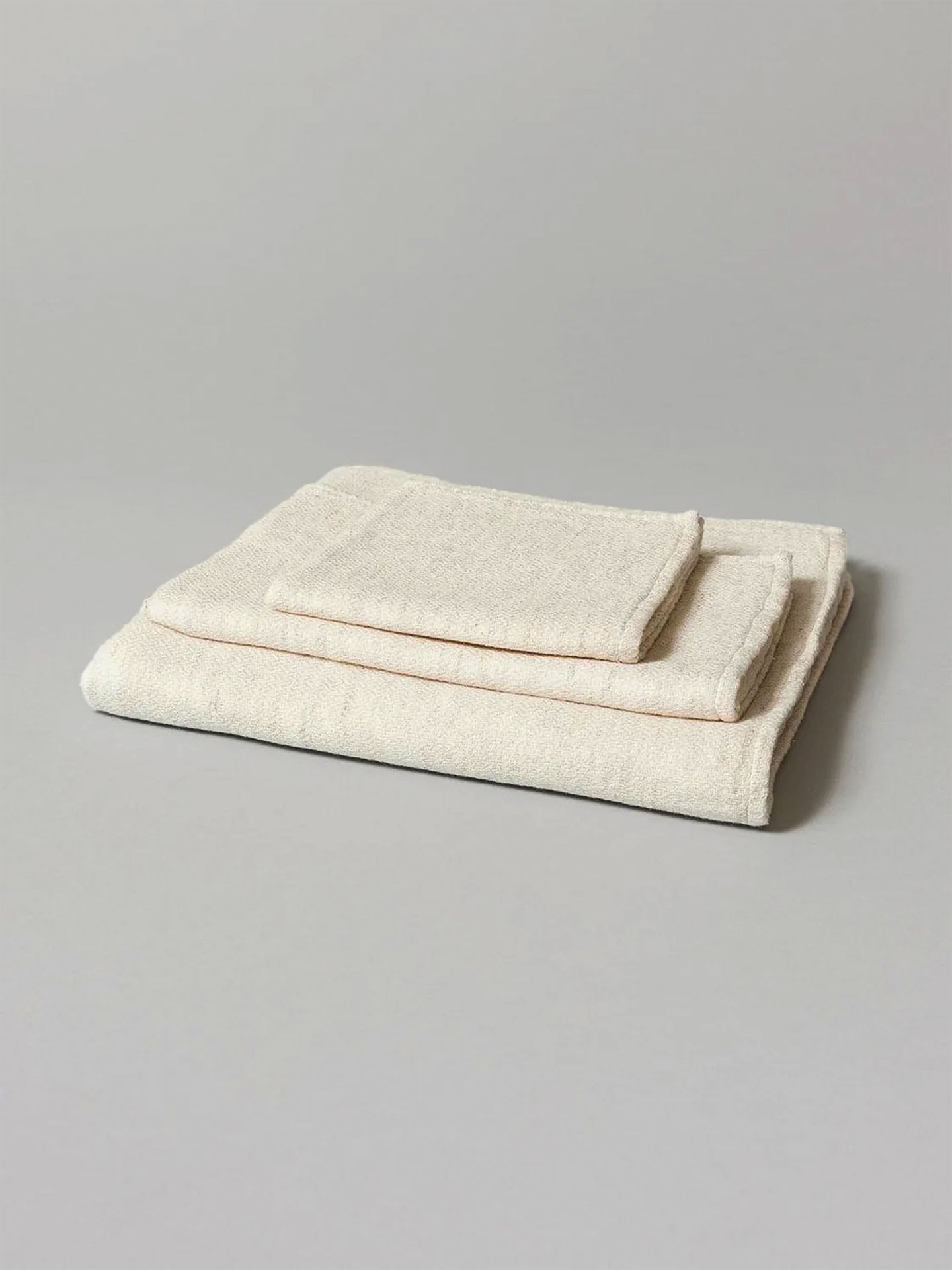 Two neatly folded Claire towels from Kontex, crafted from organic cotton and featuring one large bath towel with a smaller fast-drying hand towel on top, rest on a plain gray surface.