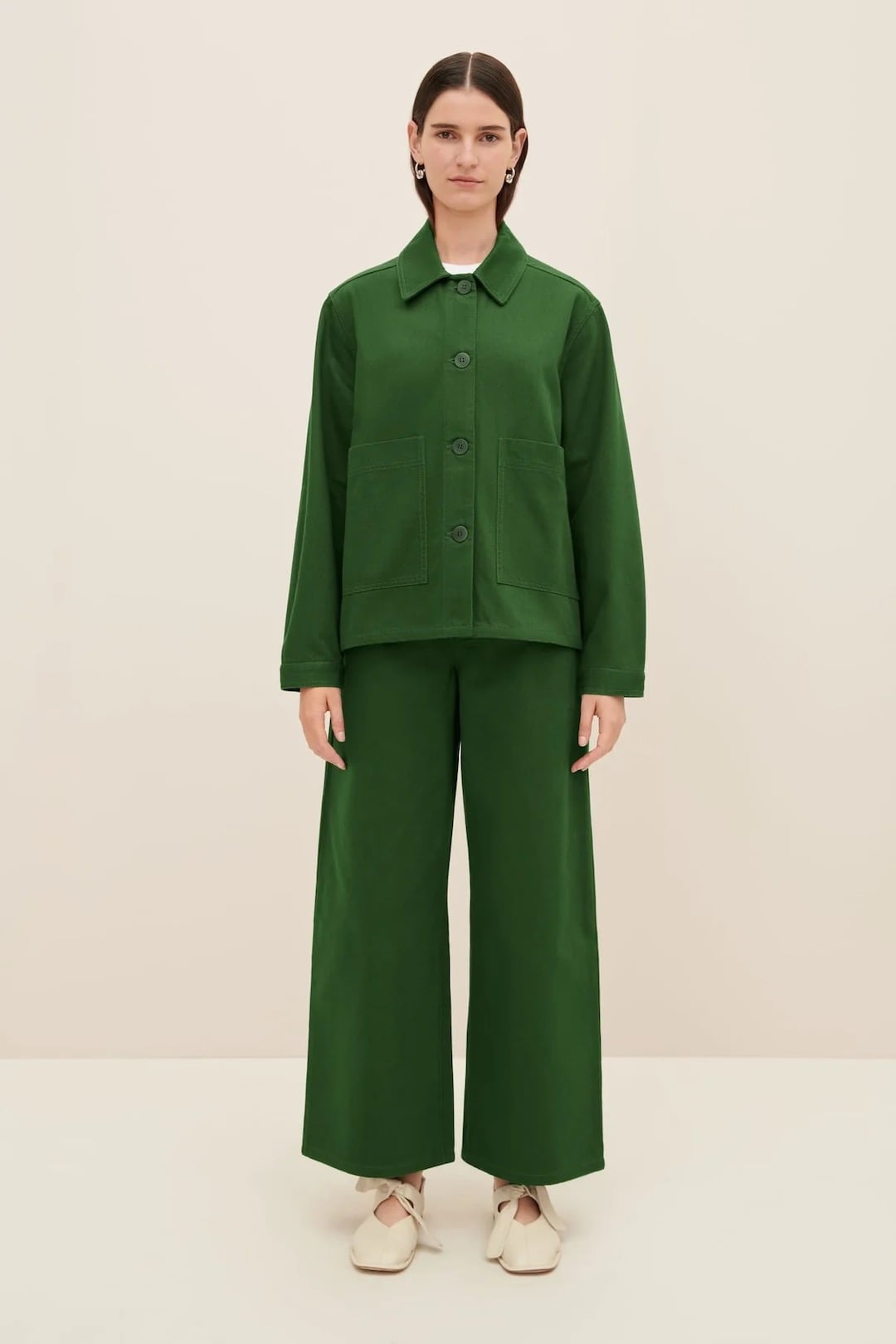 A person stands wearing the Felix Jacket in green denim by Kowtow, along with wide-legged green pants. The outfit showcases a relaxed fit that stays true to size and is complemented by white shoes against a plain background.