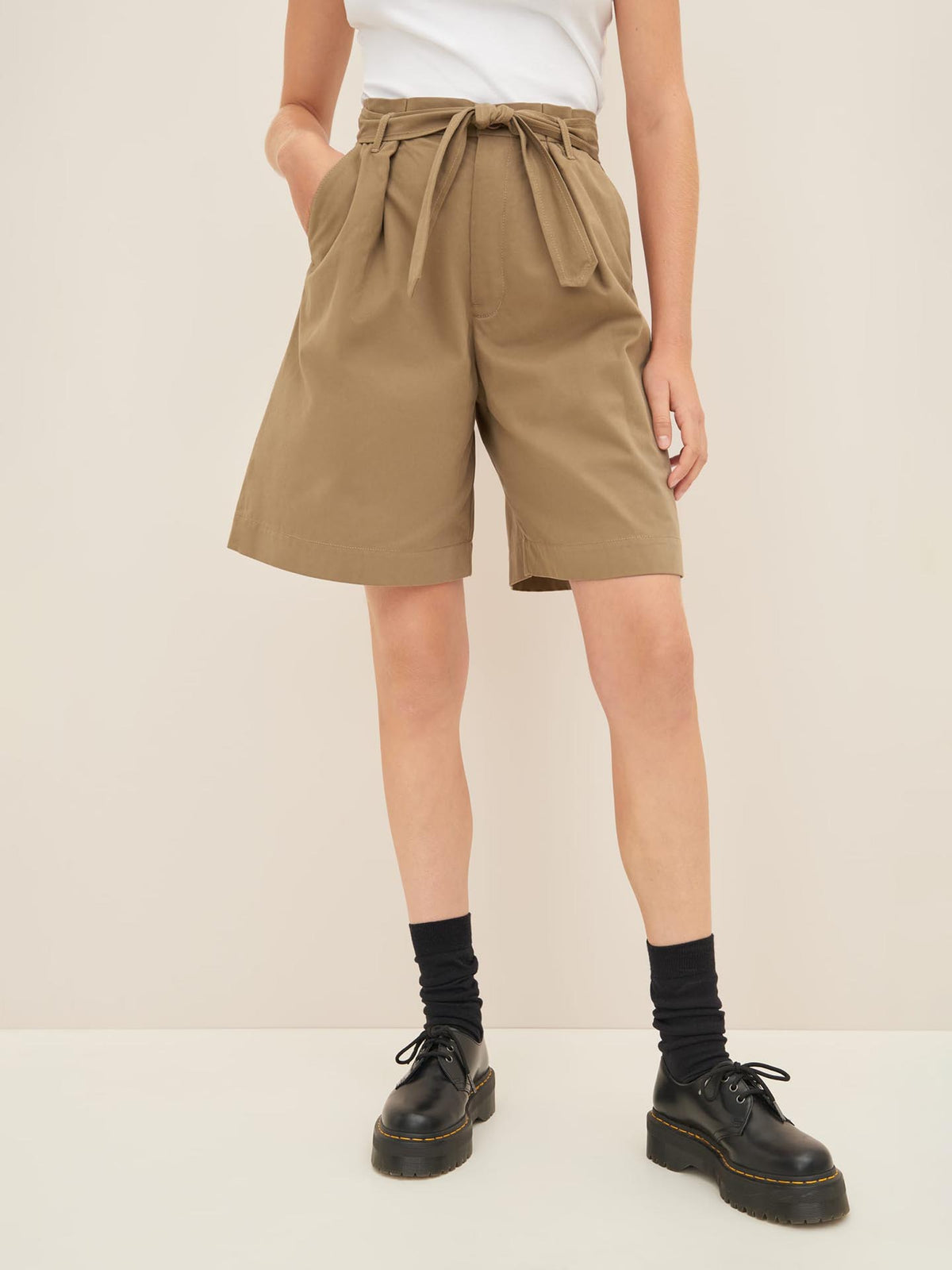 A person stands against a beige background wearing a white top, high-waisted Kowtow Alp Shorts in Bronze with a tied belt, black socks, and black shoes. The ensemble highlights the high waist design and adheres to a standard fit for easy reference in any size guide.