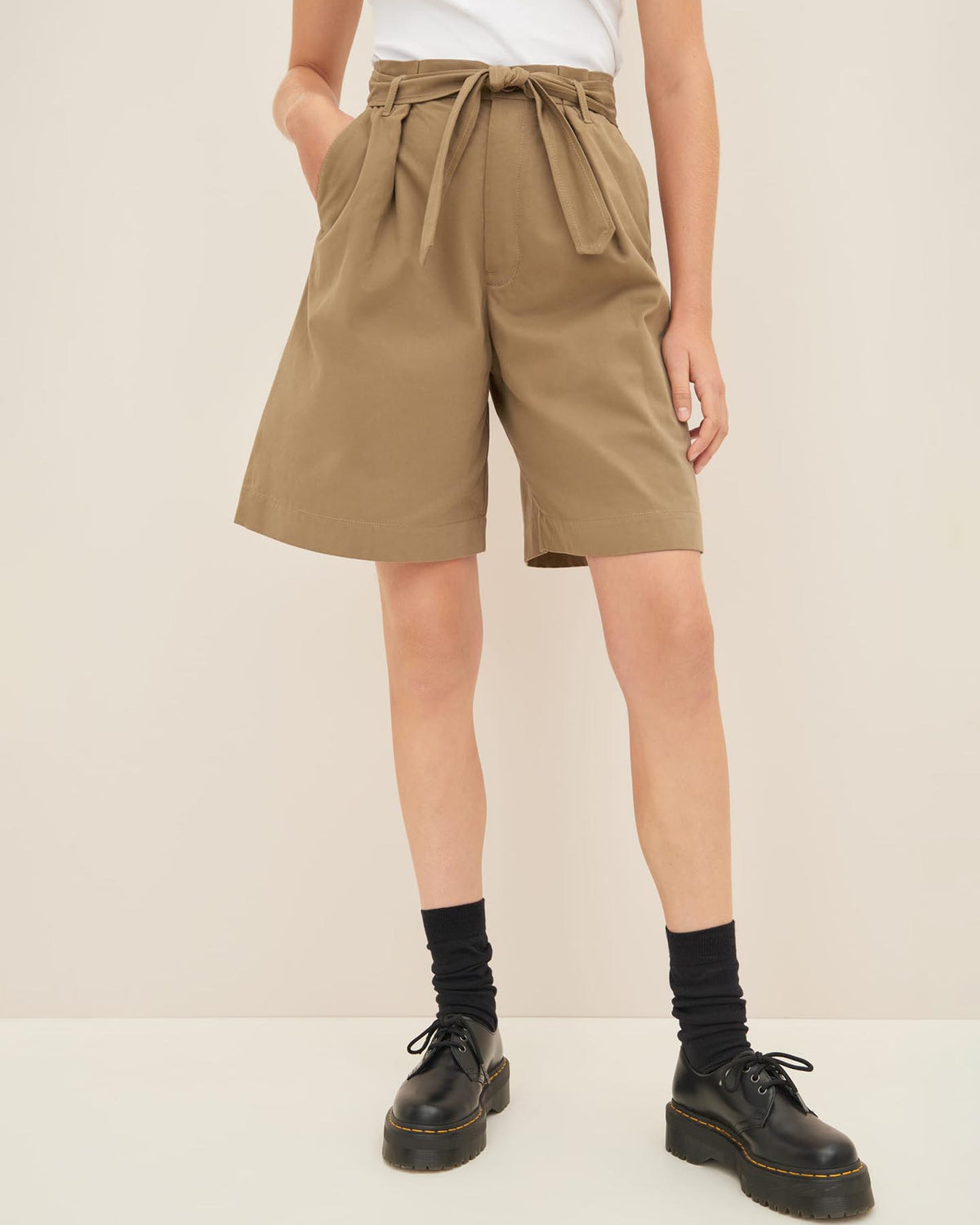 A person stands against a beige background wearing a white top, high-waisted Kowtow Alp Shorts in Bronze with a tied belt, black socks, and black shoes. The ensemble highlights the high waist design and adheres to a standard fit for easy reference in any size guide.