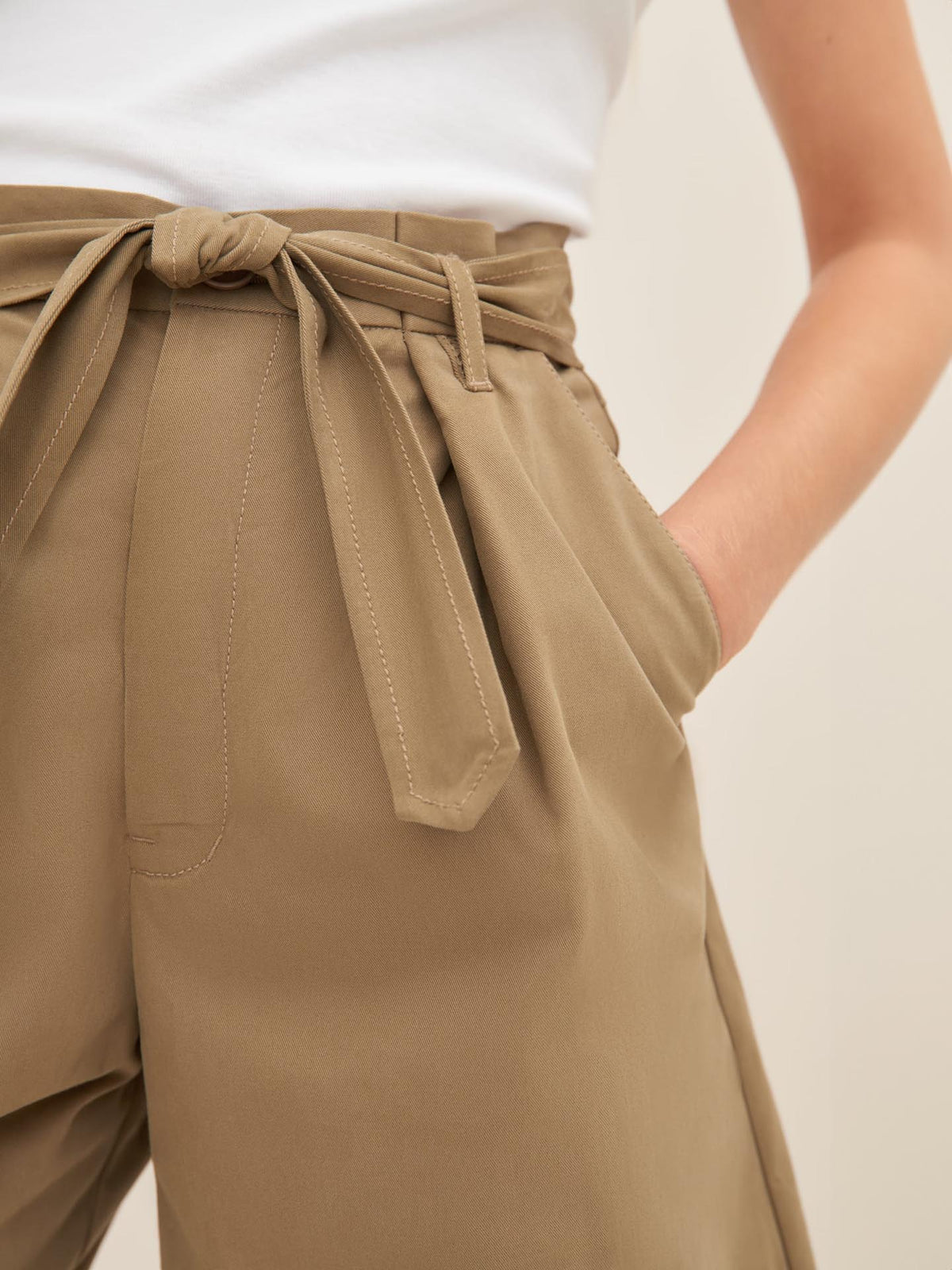 A person wearing a white shirt and Kowtow&#39;s Alp Shorts in bronze, which feature a high-waisted design with a belt tied in a bow, has their hand in one of the pockets. Check the size guide to ensure the perfect standard fit.