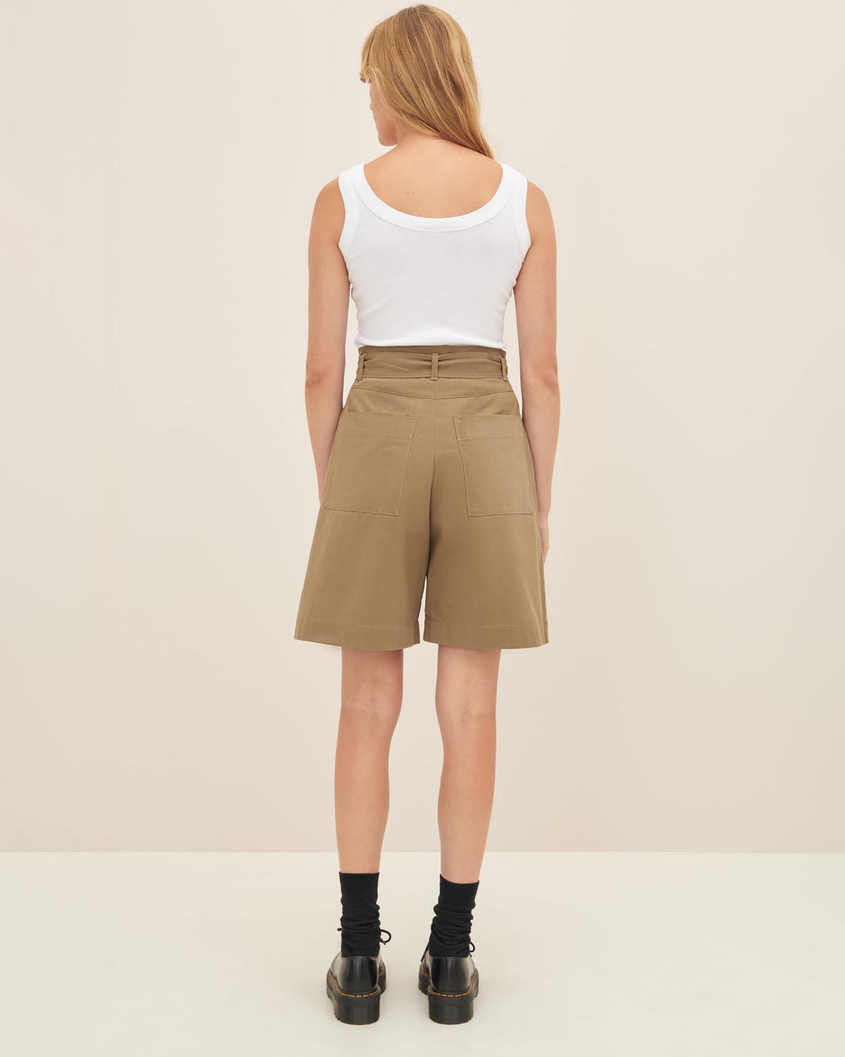 A person with long hair is standing facing away, wearing a white sleeveless top, Kowtow's Alp Shorts in Bronze, black socks, and black shoes.