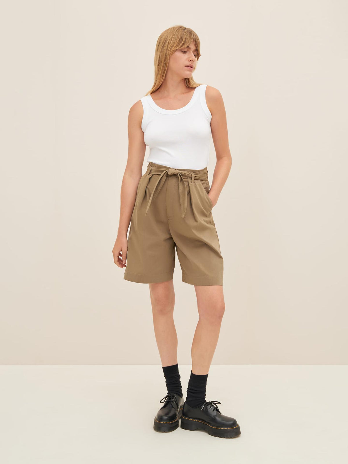 A person wearing a white sleeveless top, bronze Alp Shorts by Kowtow with a belt, black socks, and black shoes stands against a plain background, showcasing a standard fit as per the size guide.