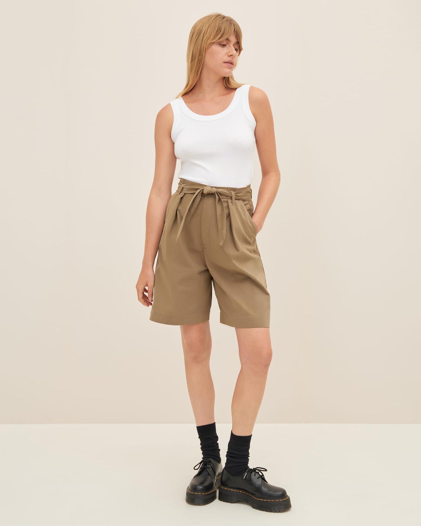 A person wearing a white sleeveless top, bronze Alp Shorts by Kowtow with a belt, black socks, and black shoes stands against a plain background, showcasing a standard fit as per the size guide.