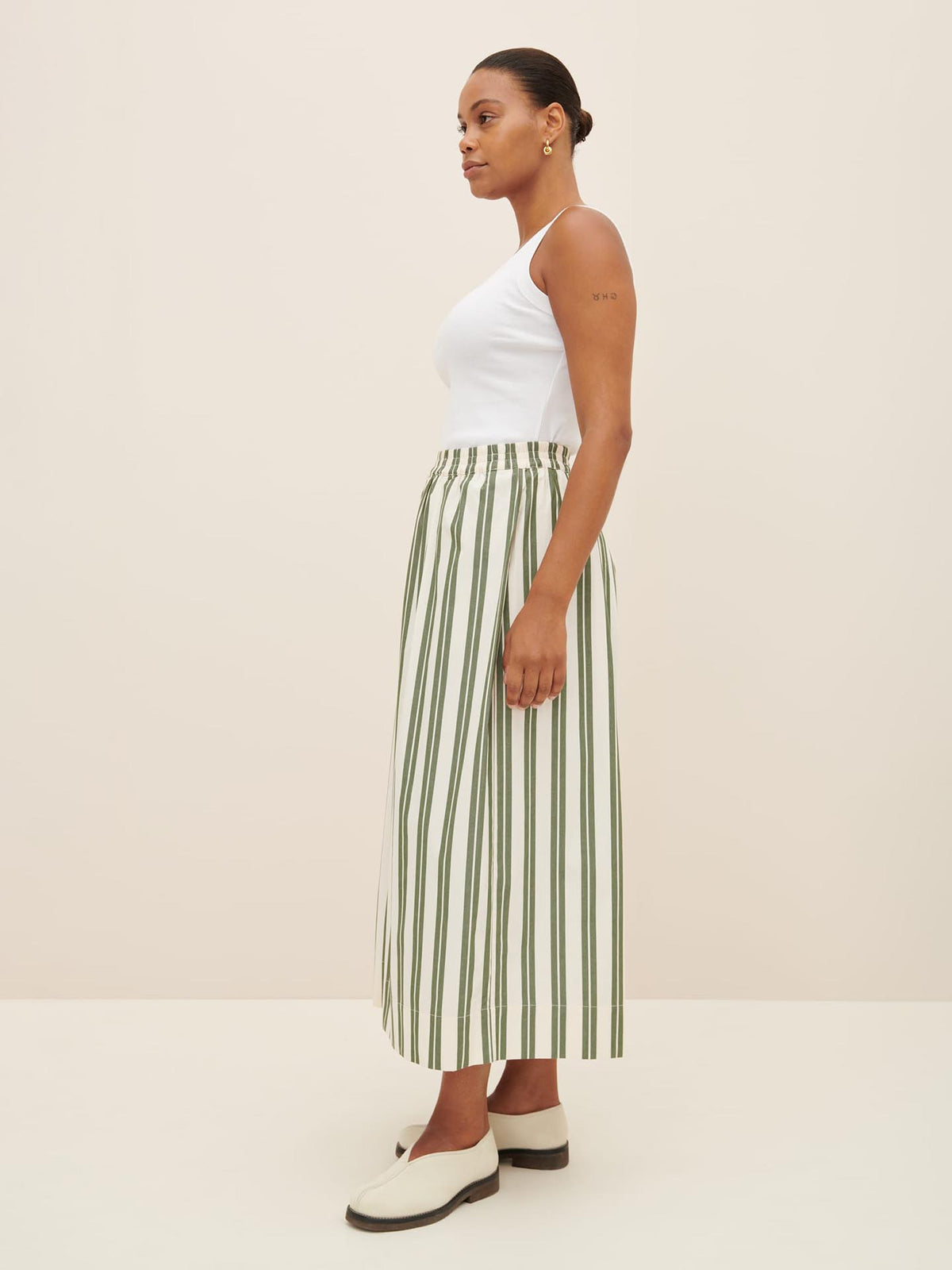 A woman in a relaxed fit white tank top, the Athena Pant in Forest Stripe by Kowtow, and white shoes stands in a neutral-colored room, facing sideways.