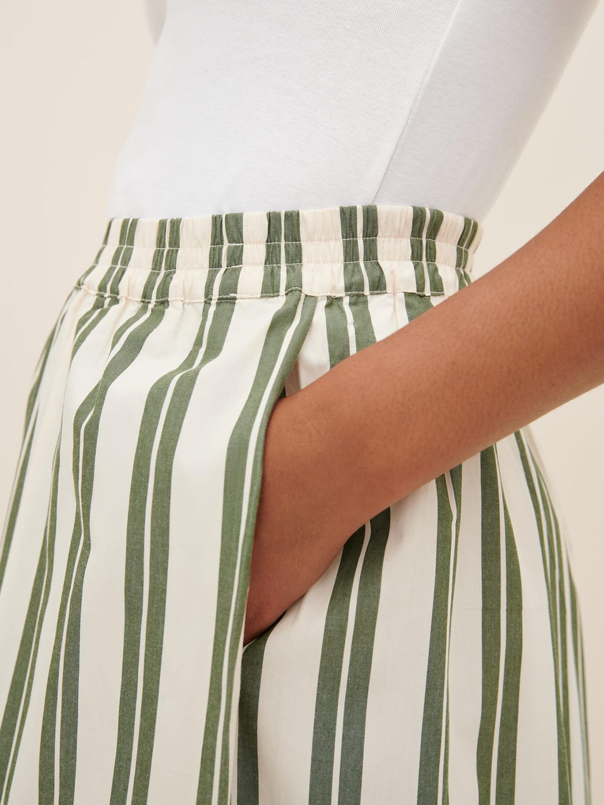 Close-up of a person wearing a white top and the Athena Pant – Forest Stripe by Kowtow, featuring high-waisted, green and white vertically striped pants with a relaxed fit, their right hand in the pant&#39;s pocket. Be sure to check the size guide if you prefer to size down for a more tailored look.