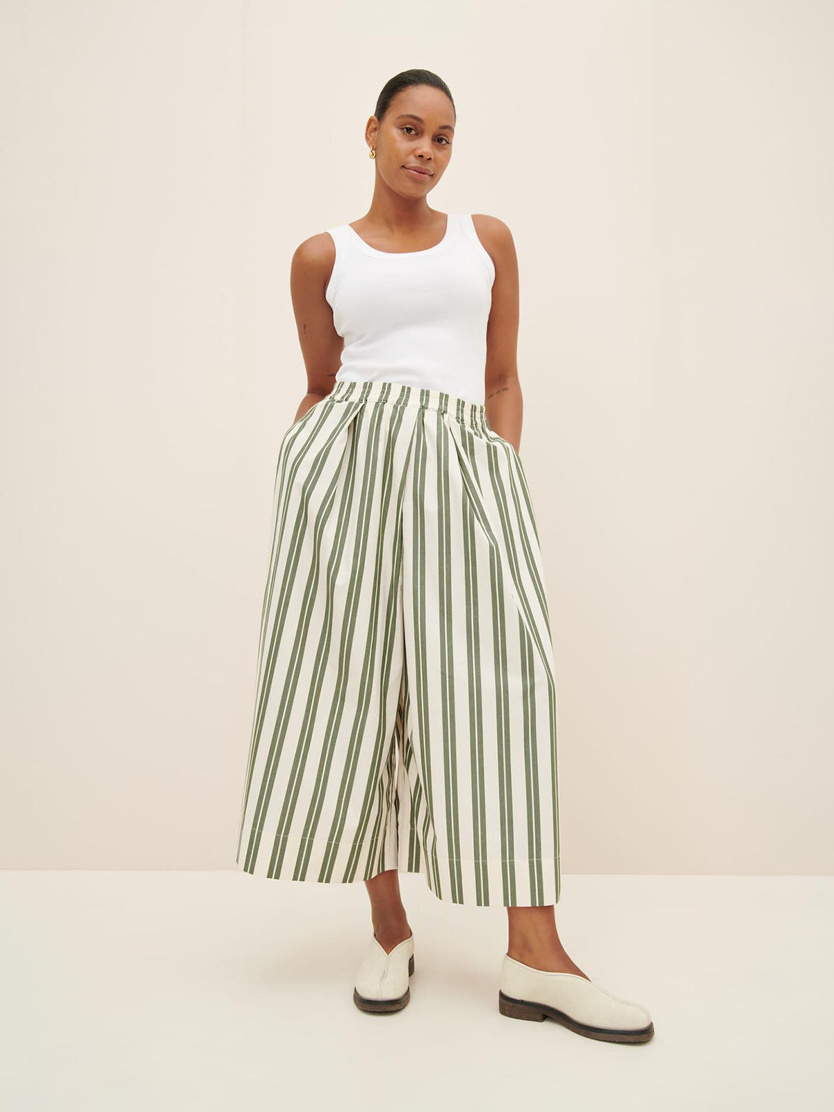 A person stands against a plain backdrop, wearing a white sleeveless top paired with the Kowtow Athena Pant in Forest Stripe. Complementing the ensemble are white slip-on shoes with black soles. The relaxed fit is accentuated by their hands casually placed in their pockets.