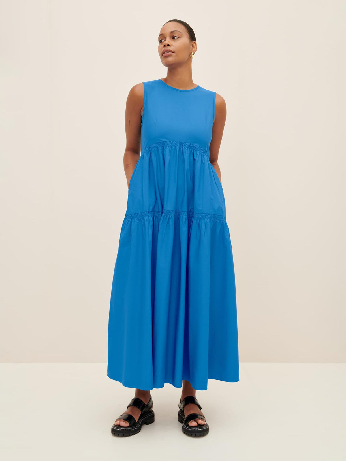 A person stands wearing the Aurora Dress, a sleeveless and long blue gown in Kowtow size XS, paired with black sandals against a plain background.