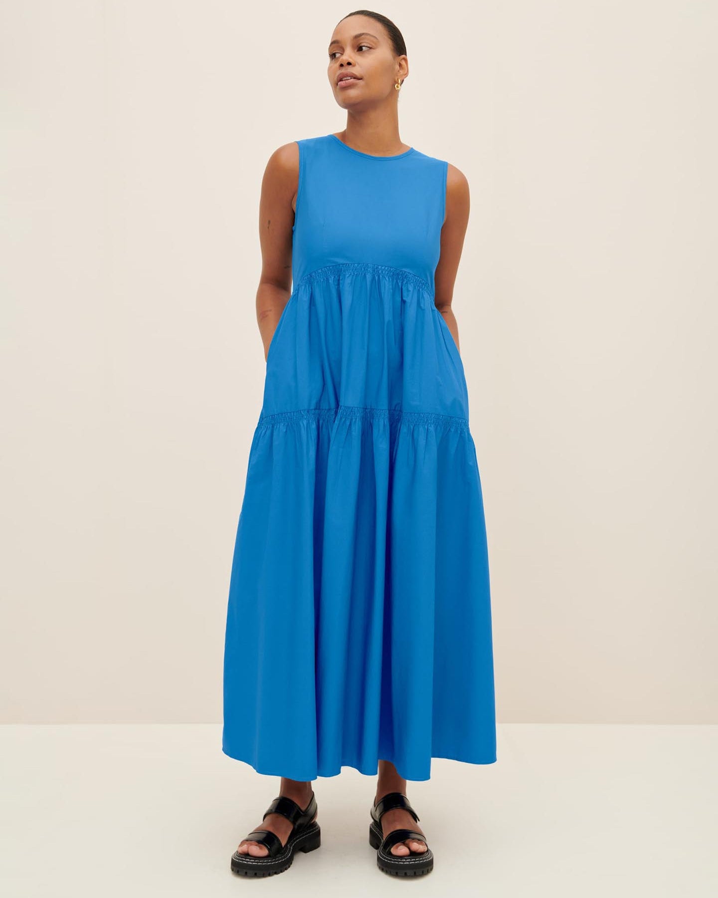 A person stands wearing the Aurora Dress, a sleeveless and long blue gown in Kowtow size XS, paired with black sandals against a plain background.