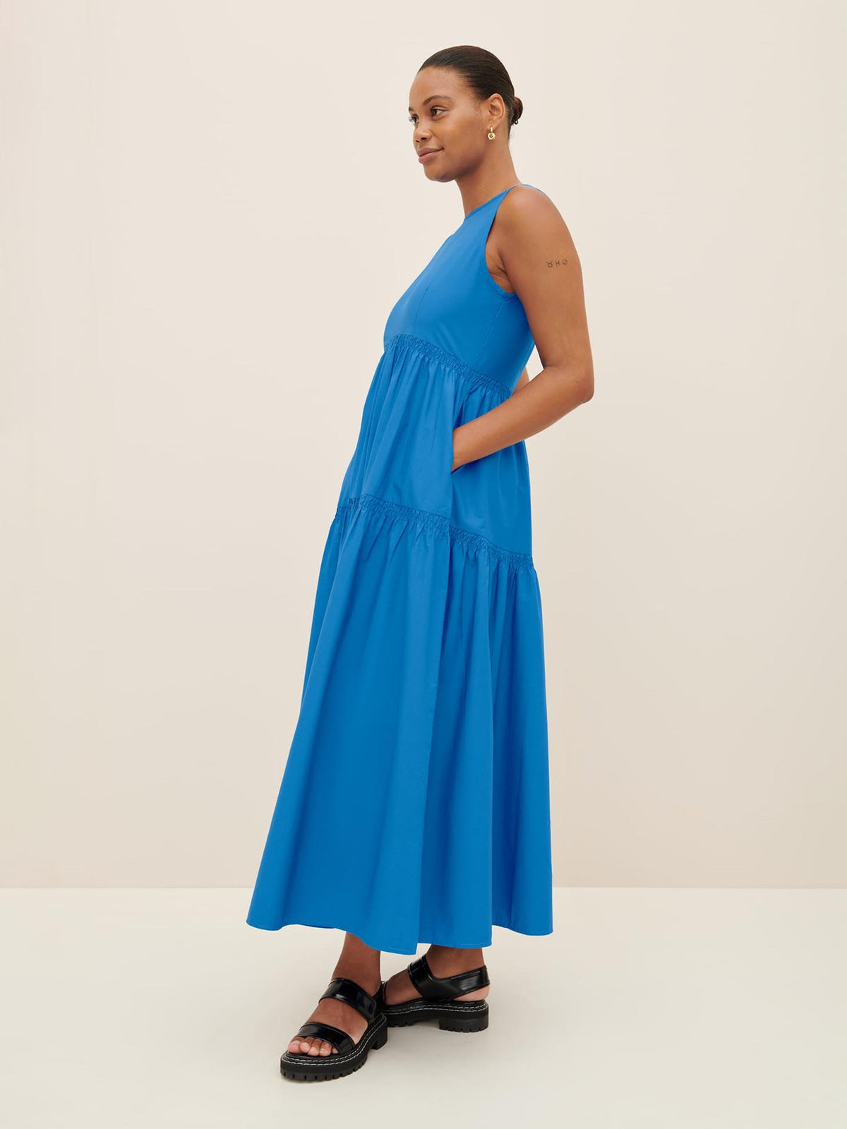 A person is elegantly styled in the Kowtow Aurora Dress, a sleeveless maxi dress in blue, paired with black sandals. They pose gracefully against a plain backdrop, showcasing the sophistication of the size XS collection.