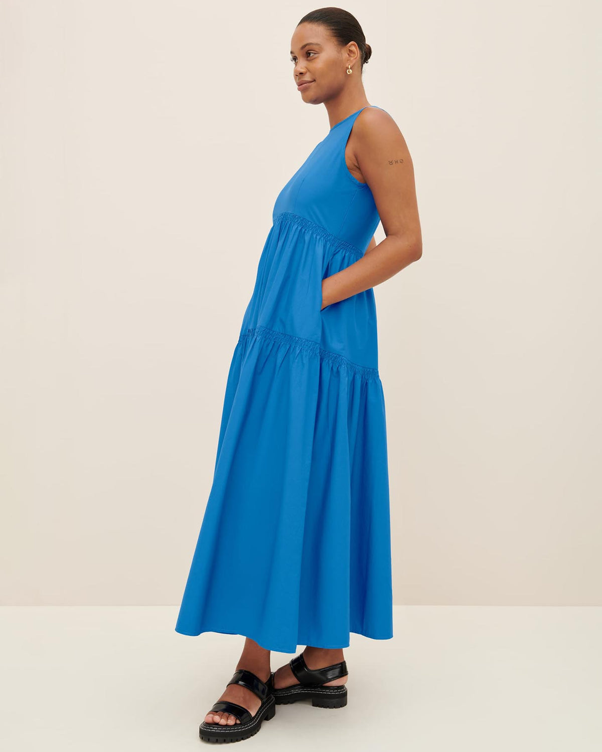 A person is elegantly styled in the Kowtow Aurora Dress, a sleeveless maxi dress in blue, paired with black sandals. They pose gracefully against a plain backdrop, showcasing the sophistication of the size XS collection.