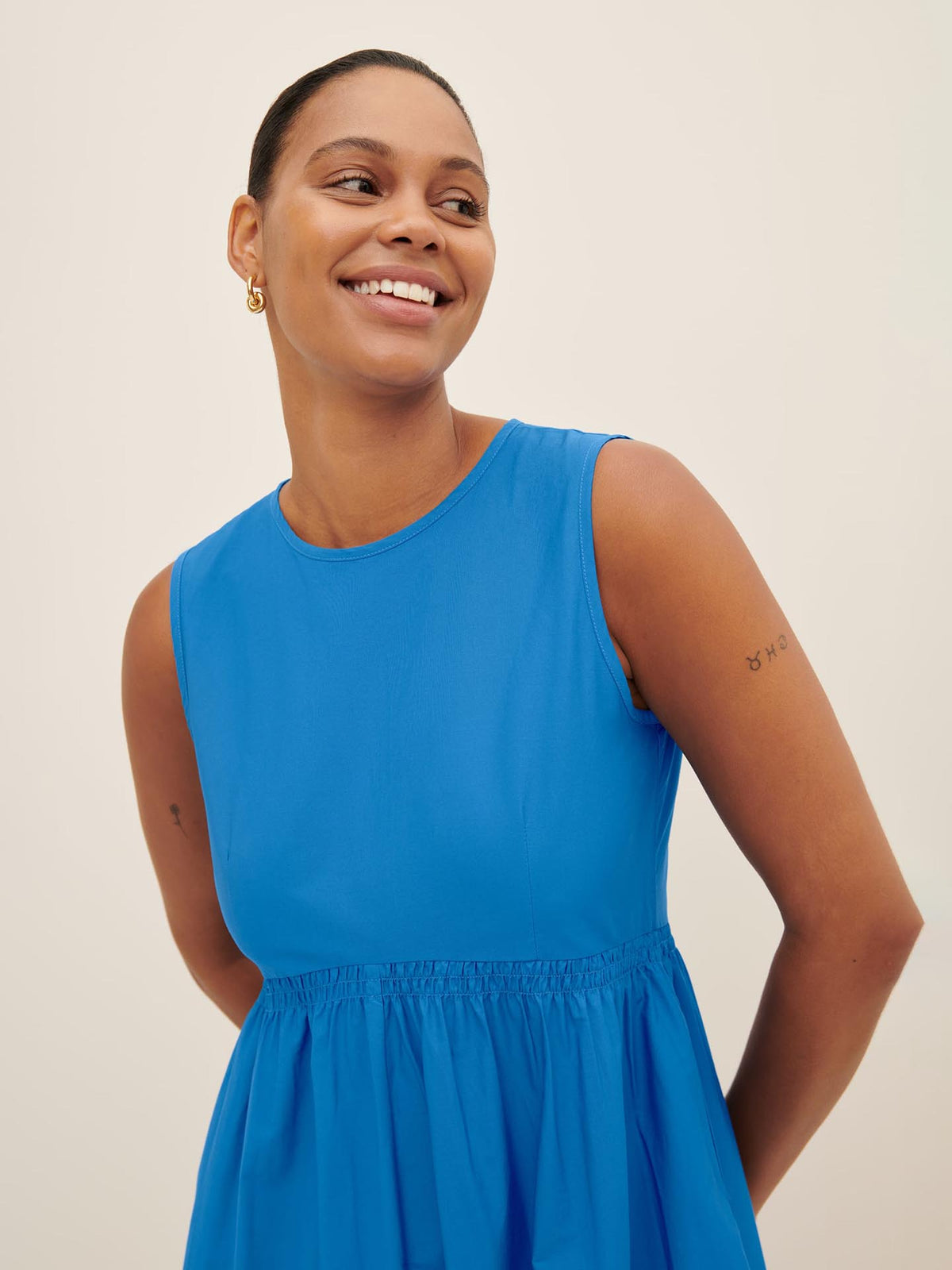 A person smiles in the Kowtow Aurora Dress, a stylish sleeveless blue piece that artfully displays small tattoos on both arms as she poses with her hands behind her back. This dress provides a standard fit akin to Kowtow size XS, making it an excellent choice for individuals prioritizing both comfort and style. Refer to our size guide to find your perfect fit.