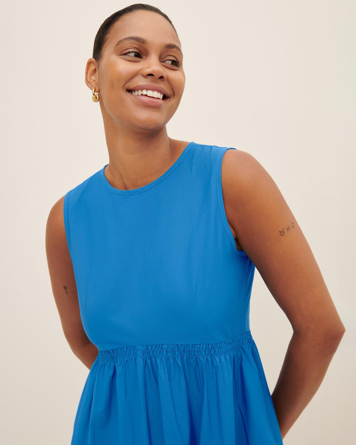 A person smiles in the Kowtow Aurora Dress, a stylish sleeveless blue piece that artfully displays small tattoos on both arms as she poses with her hands behind her back. This dress provides a standard fit akin to Kowtow size XS, making it an excellent choice for individuals prioritizing both comfort and style. Refer to our size guide to find your perfect fit.