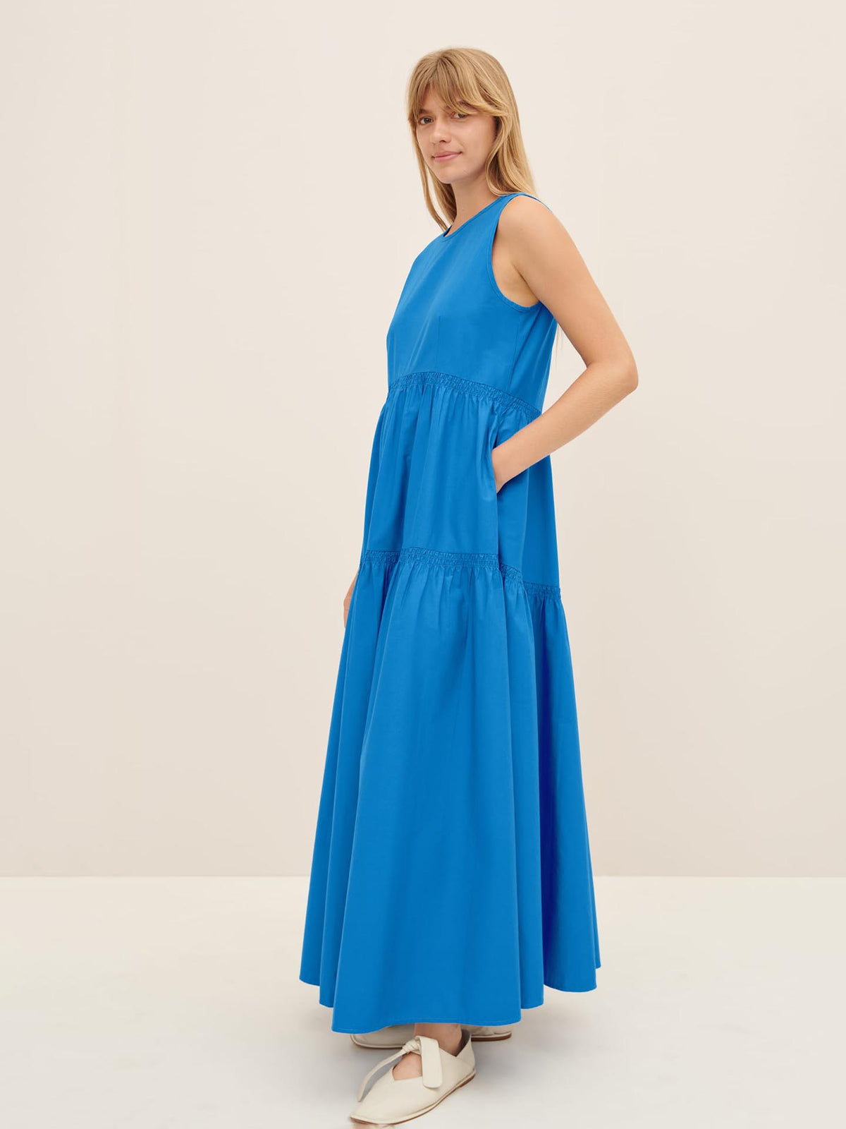 A person wearing the Aurora Dress by Kowtow, a long and sleeveless blue dress in a standard fit, stands against a plain background, perfectly sized according to the size XS guide.