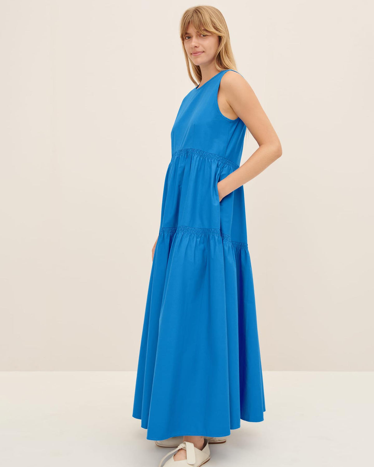 A person wearing the Aurora Dress by Kowtow, a long and sleeveless blue dress in a standard fit, stands against a plain background, perfectly sized according to the size XS guide.
