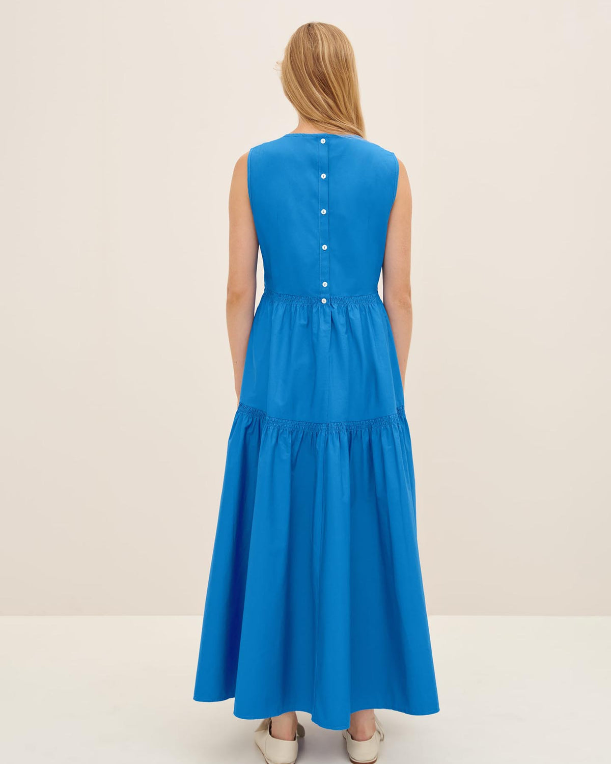 A person with long hair is wearing the Aurora Dress by Kowtow in size XS, featuring a row of buttons on the back, and is standing in a light-colored room.