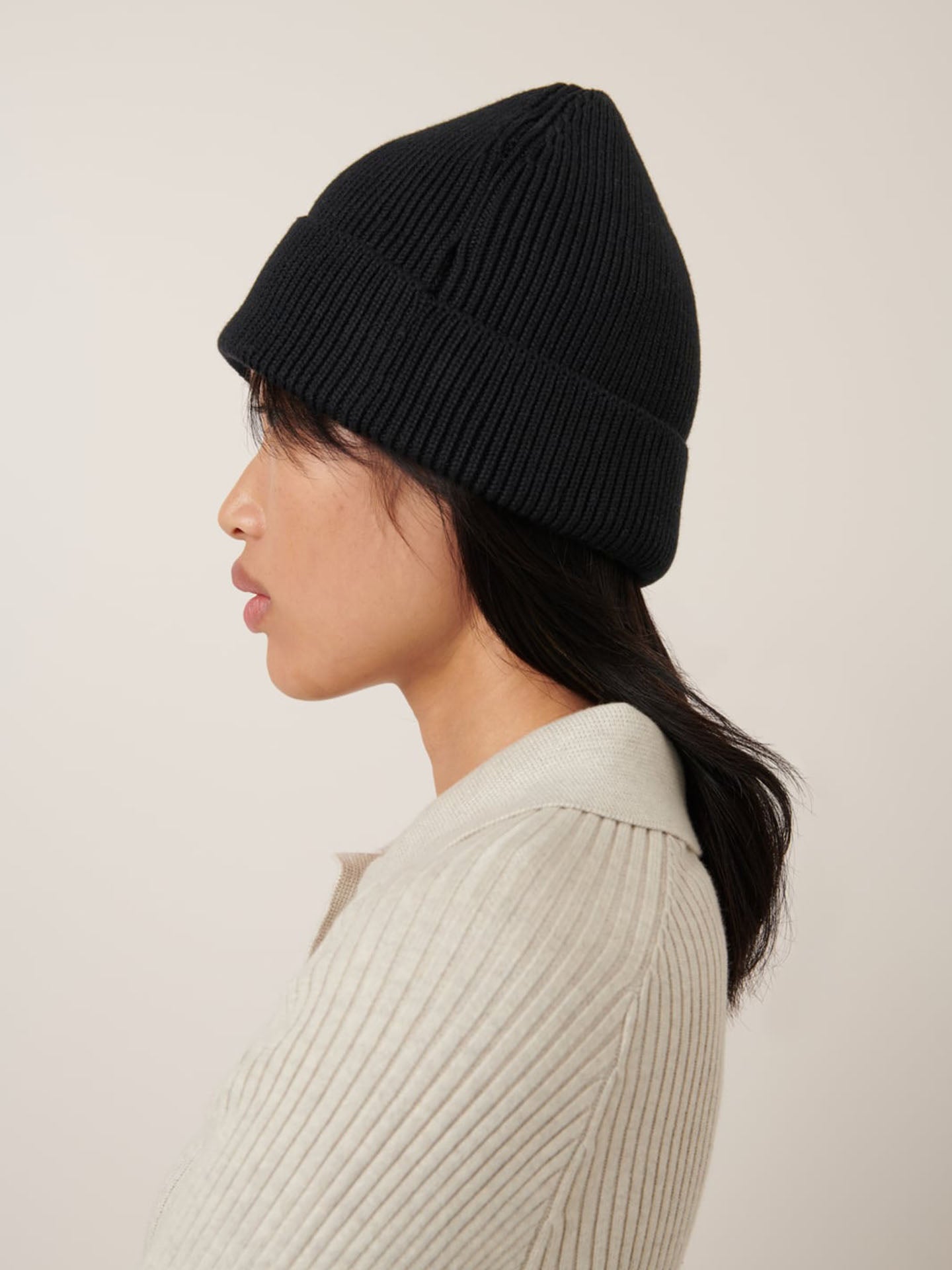 In profile, a person faces left against a plain background, wearing Kowtow's black Beanie with a rolled cuff and a light ribbed sweater for a comfortable fit.