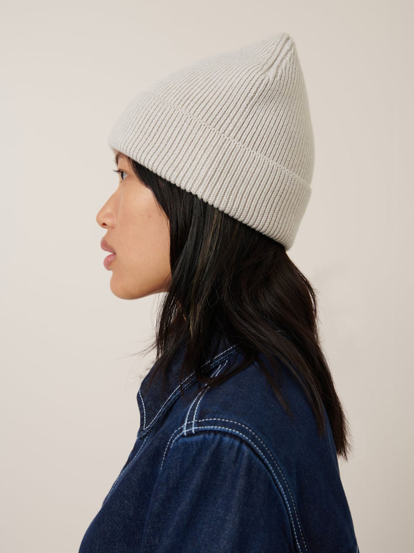 A person in profile wears the Kowtow Beanie – Oat Melange, featuring a snug fit and rolled cuff, paired with a dark denim jacket, facing left against a plain backdrop.