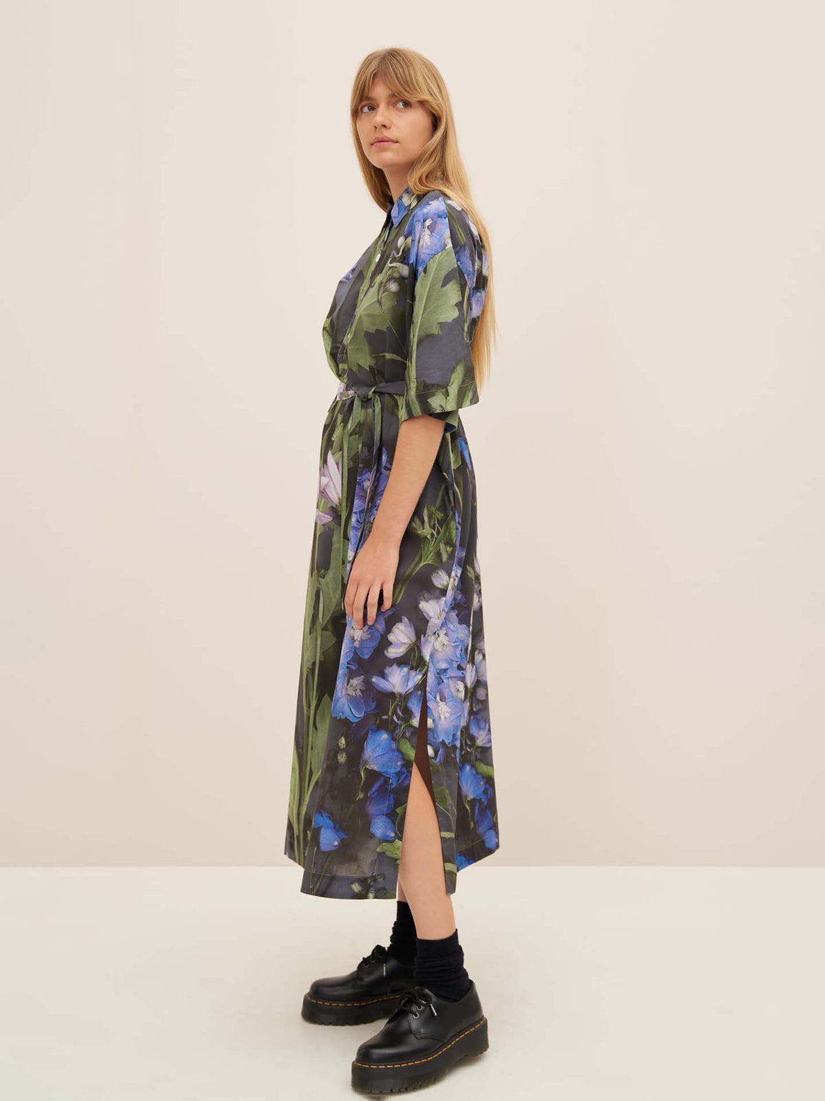 A woman with long hair wears the Bloom Dress by Kowtow, featuring a floral print and a standard fit, paired with black socks and platform shoes. She stands against a plain background, embodying elegance effortlessly.