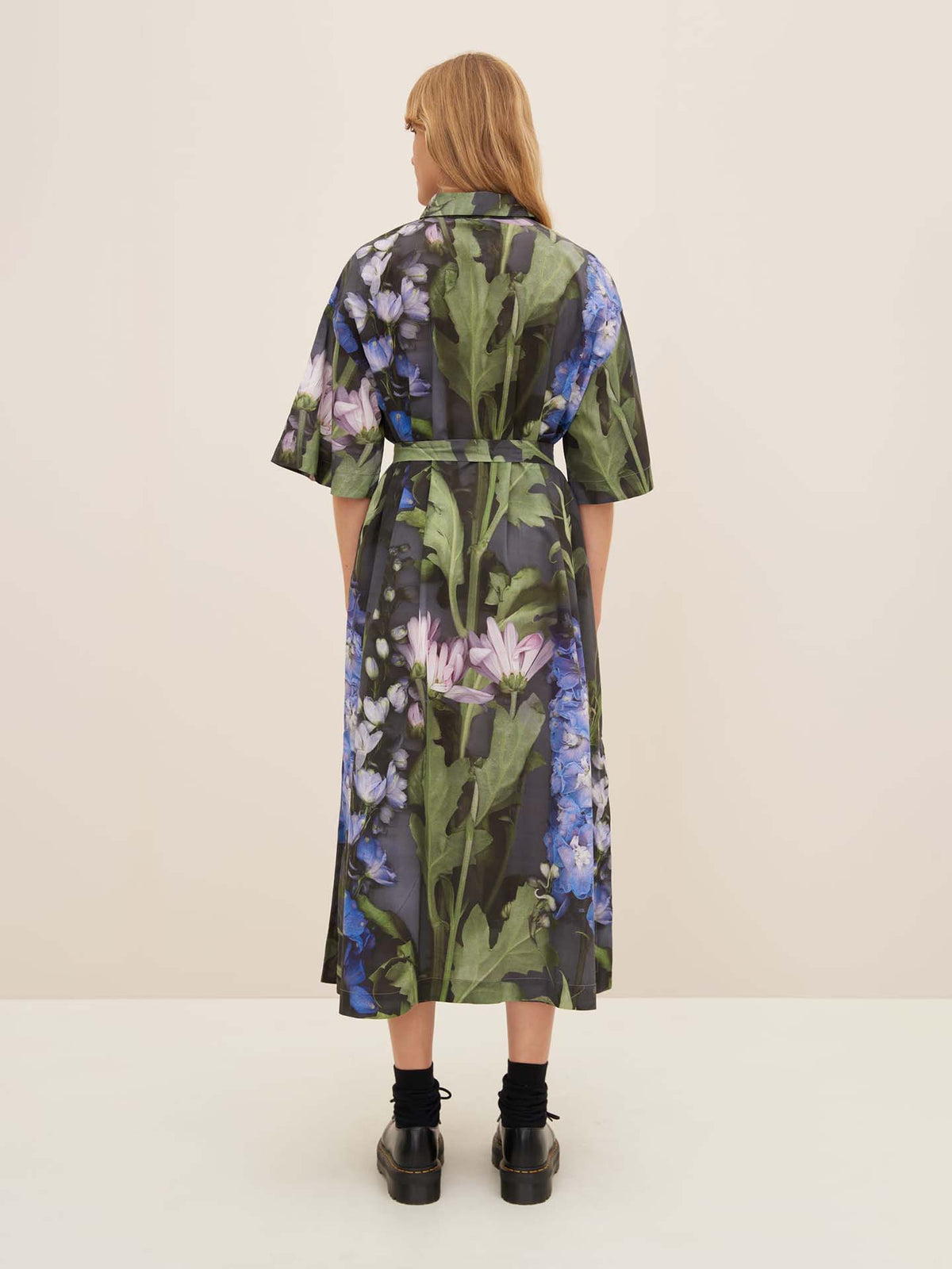 A person is shown from the back wearing a Kowtow Bloom Dress, featuring a floral pattern and short sleeves, while standing on a neutral background.