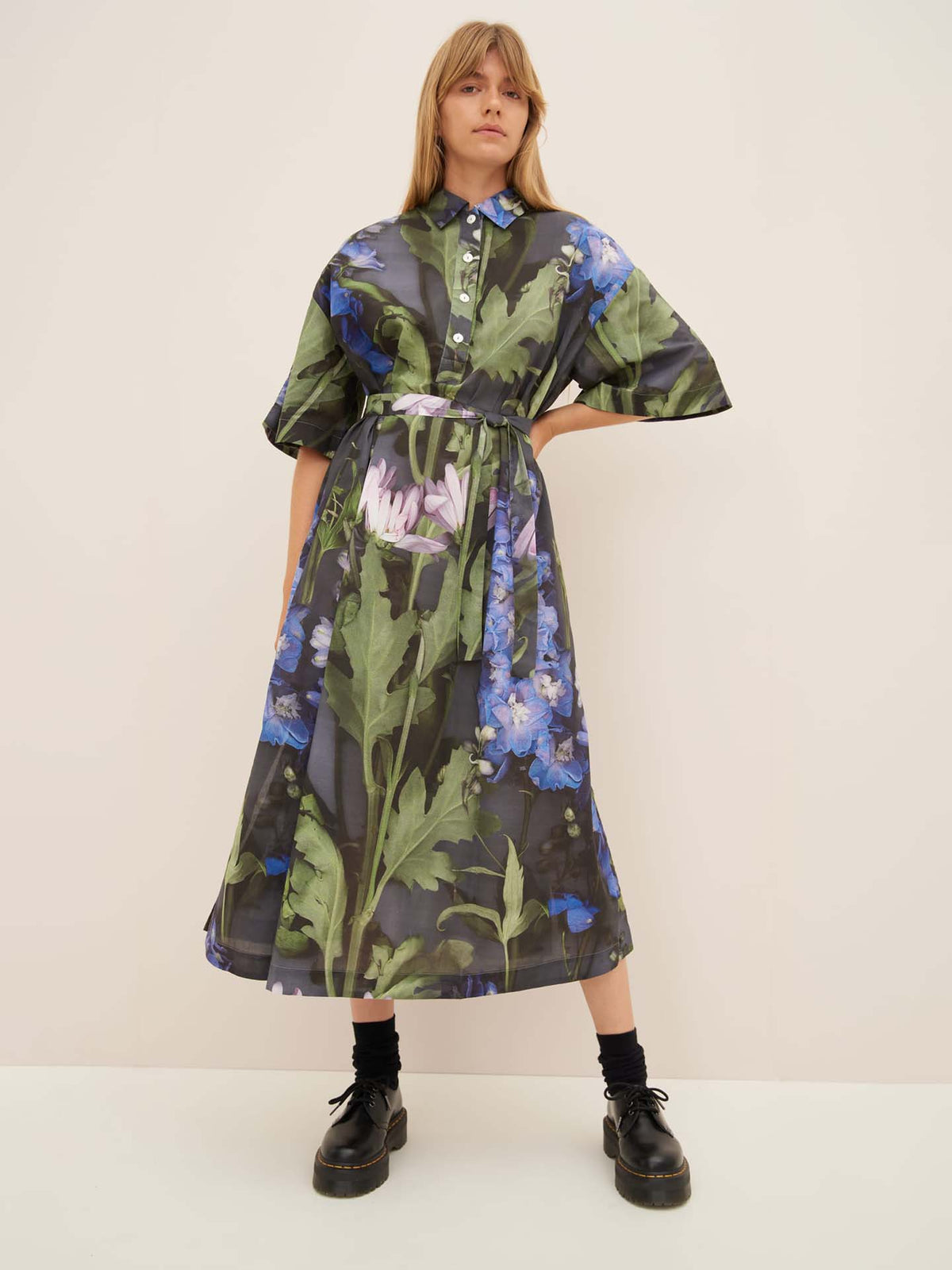 A person is wearing the Bloom Dress by Kowtow, showcasing its standard fit with blue, purple, and green floral patterns. The look is completed with black shoes and socks against a neutral background as they stand with hands on hips.