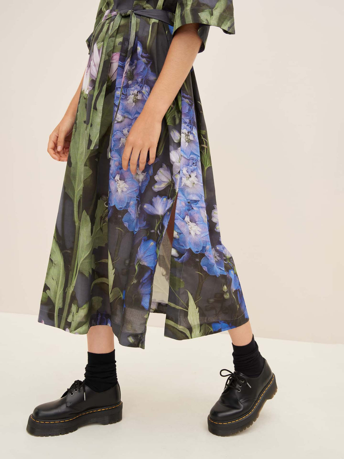 Wearing the Bloom Dress by Kowtow, adorned with a stunning floral pattern of blue and purple flowers, along with black socks and chunky black shoes, a person confidently embodies the essence of standard fit elegance.