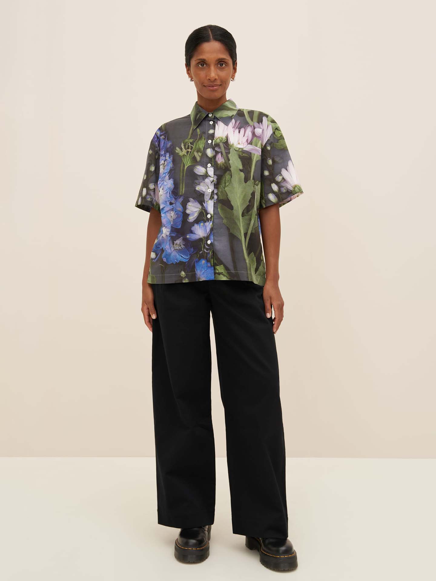 A person wearing the Bloom Shirt by Kowtow, a floral button-up with a relaxed fit and dropped shoulder seams, paired with black pants, stands in a neutral pose against a plain background.