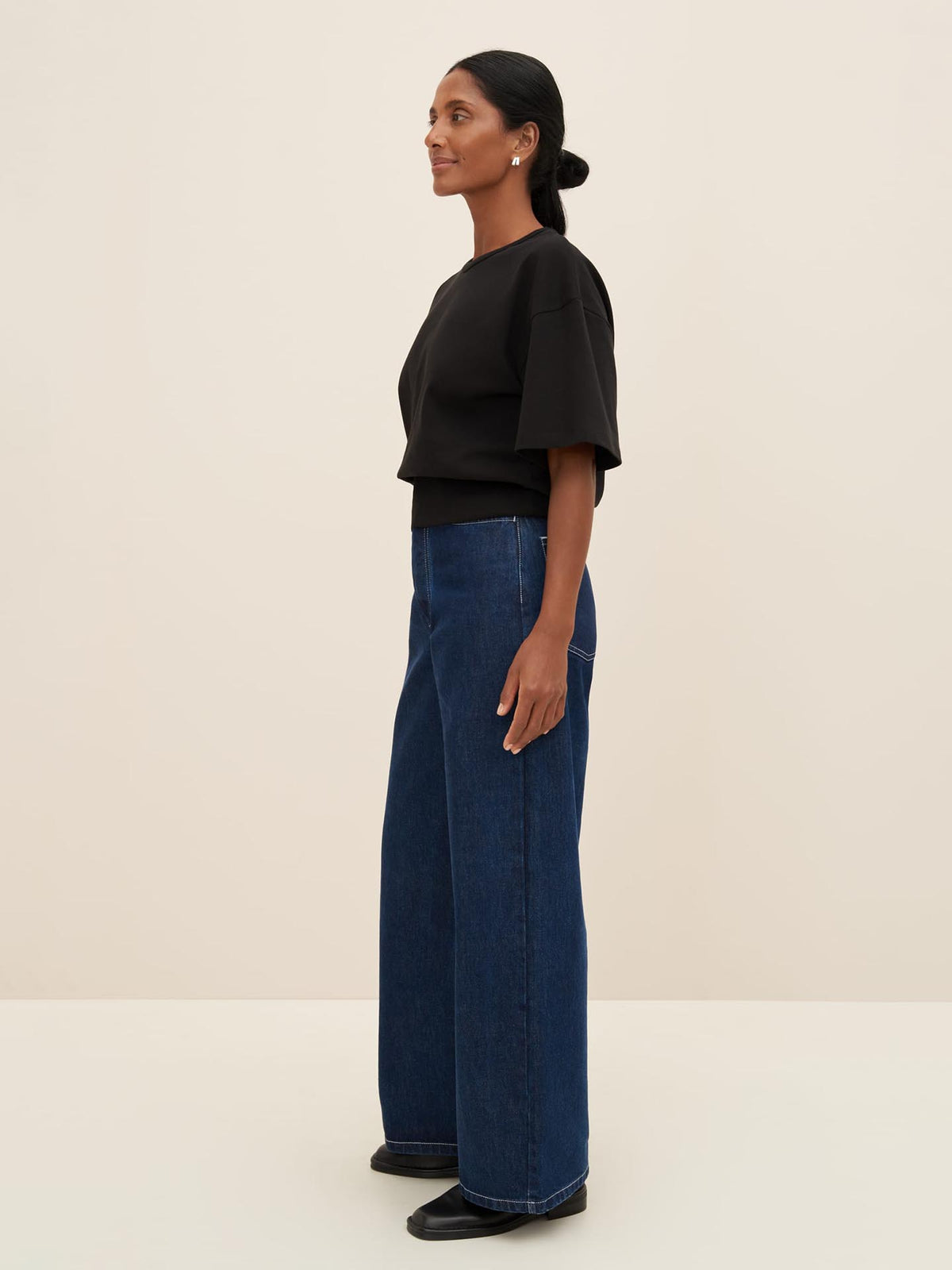 A person stands sideways, wearing a black &quot;Boxy Rib Tee&quot; from Kowtow and wide-leg blue jeans with a relaxed fit. The background is plain and neutral-colored.