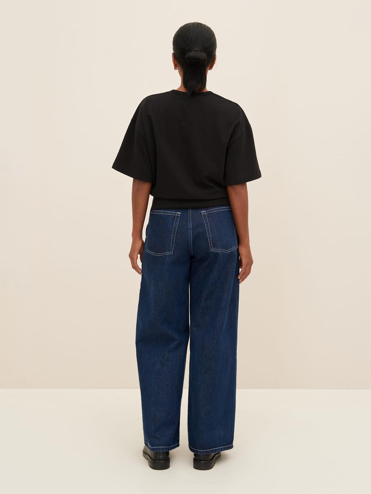 A person stands facing away from the camera, sporting a black Boxy Rib Tee from Kowtow, paired with blue wide-leg jeans that have visible back pockets and black shoes, all showcasing a relaxed fit. For sizing reference, they are wearing a Kowtow size XS. The backdrop is plain beige.