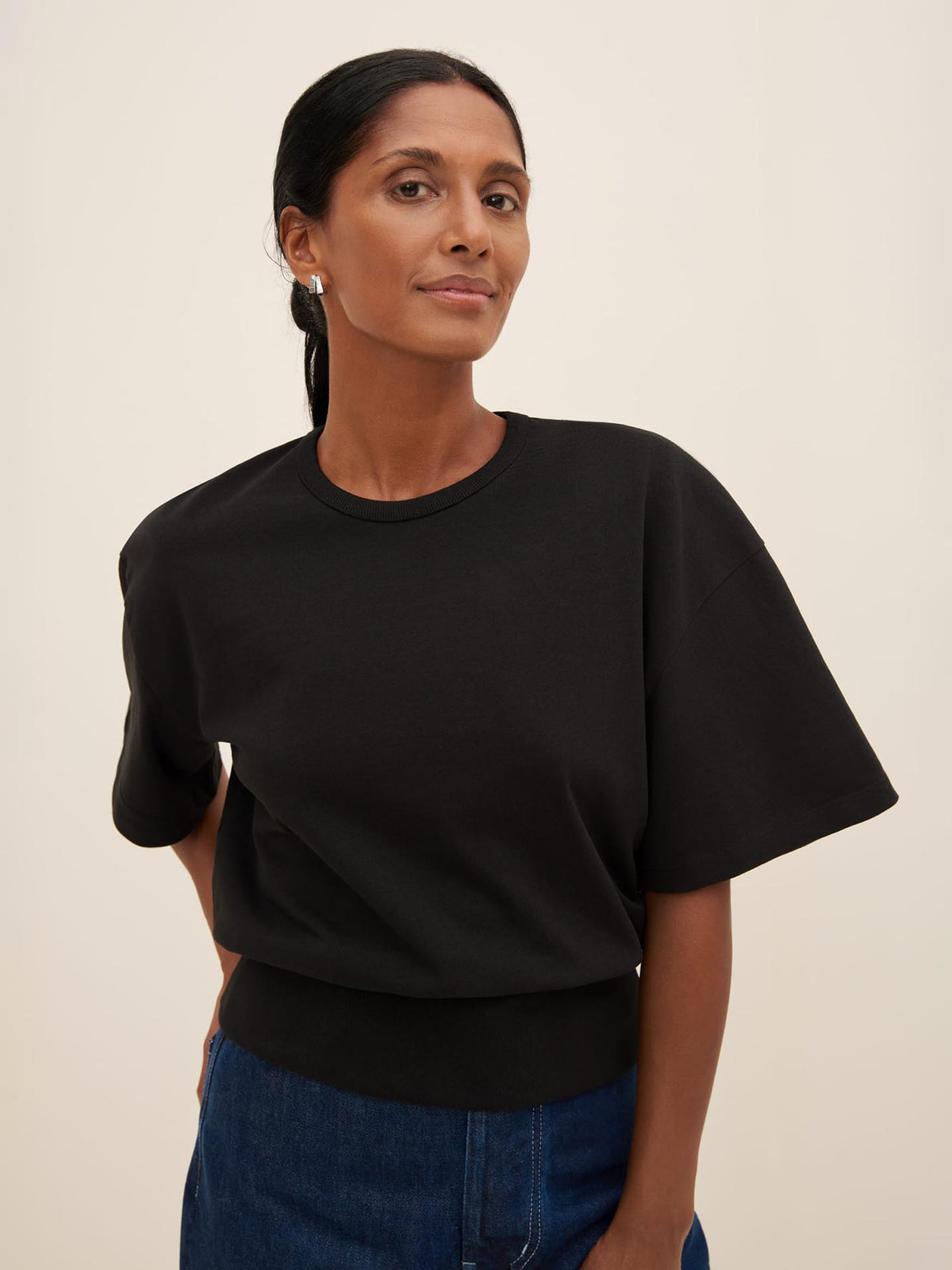 A person with dark hair pulled back wears a Kowtow Boxy Rib Tee in black and blue jeans with a relaxed fit, standing against a plain background.