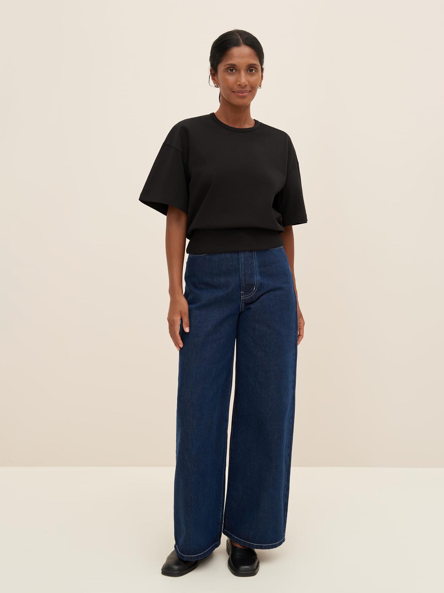 A person stands against a plain background, wearing the Kowtow Boxy Rib Tee in black, wide-leg blue jeans in size XS for a relaxed fit, and black shoes.