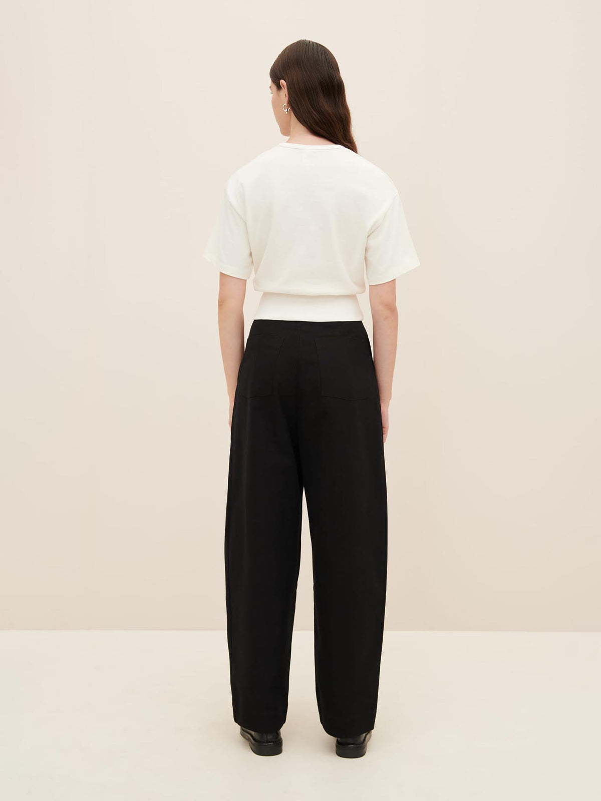 A person with long dark hair is standing with their back to the camera, wearing a white Boxy Rib Tee by Kowtow, black wide-leg pants, and black shoes, against a plain light backdrop.