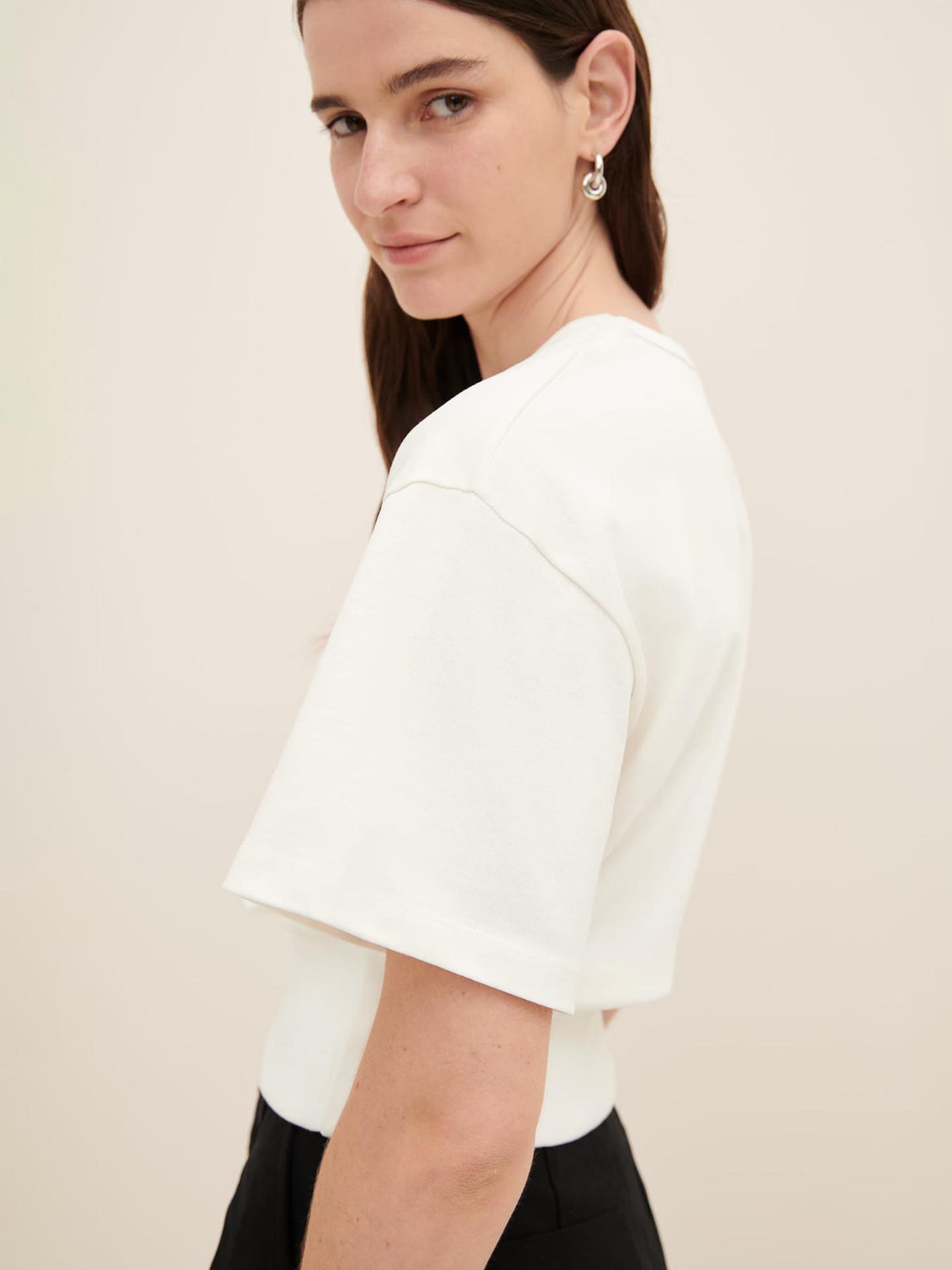 Woman with long brown hair, wearing the white Boxy Rib Tee from Kowtow and silver earrings, looking over her shoulder at the camera. The shirt has an oversized fit that enhances its casual charm.