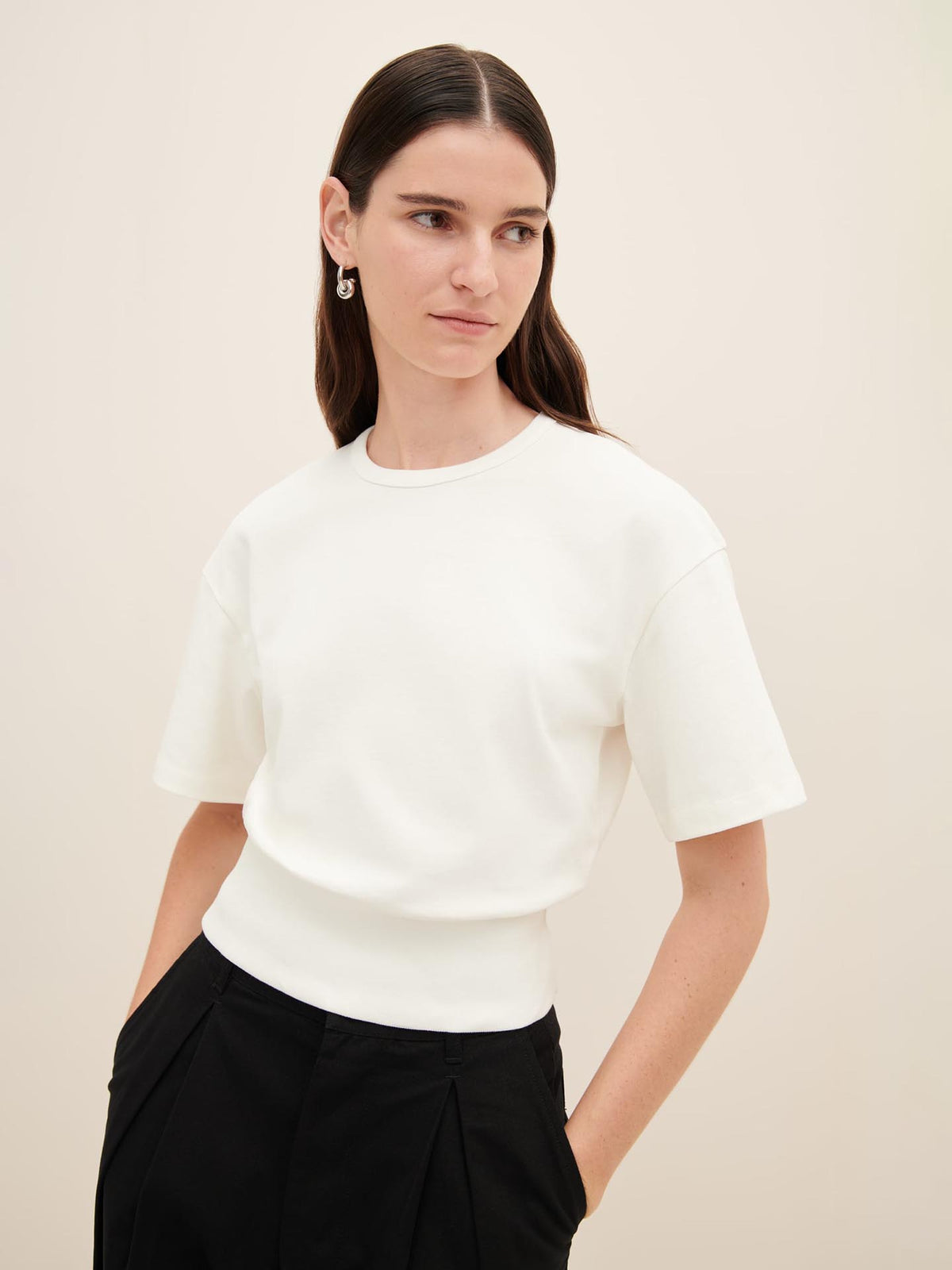 A person with long brown hair is wearing the Boxy Rib Tee – White by Kowtow and black trousers. They are standing against a plain background, looking slightly to the side.