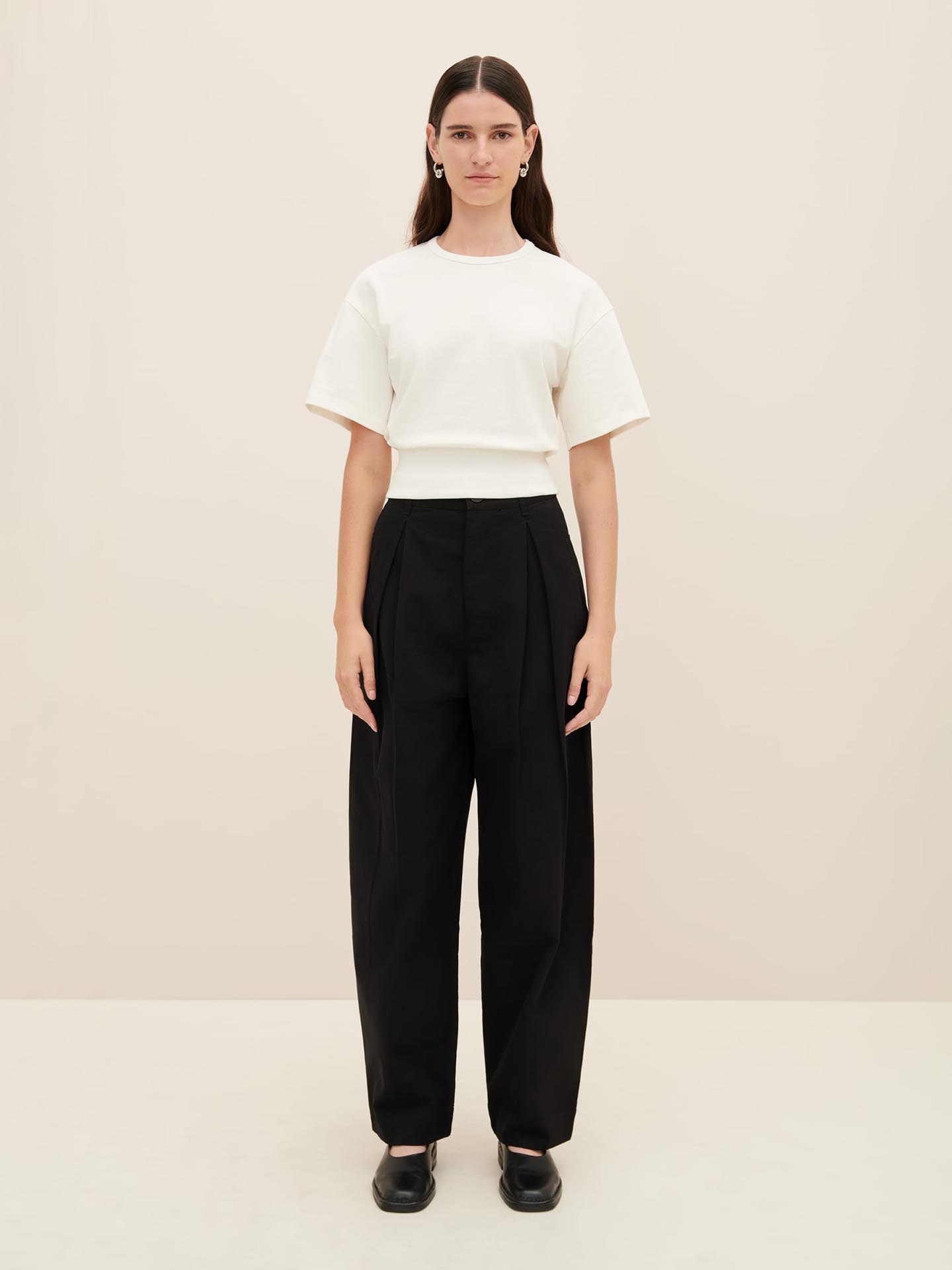 A person with long dark hair stands against a plain background, wearing the Kowtow Boxy Rib Tee in white, paired with black high-waisted wide-leg pants and black shoes.