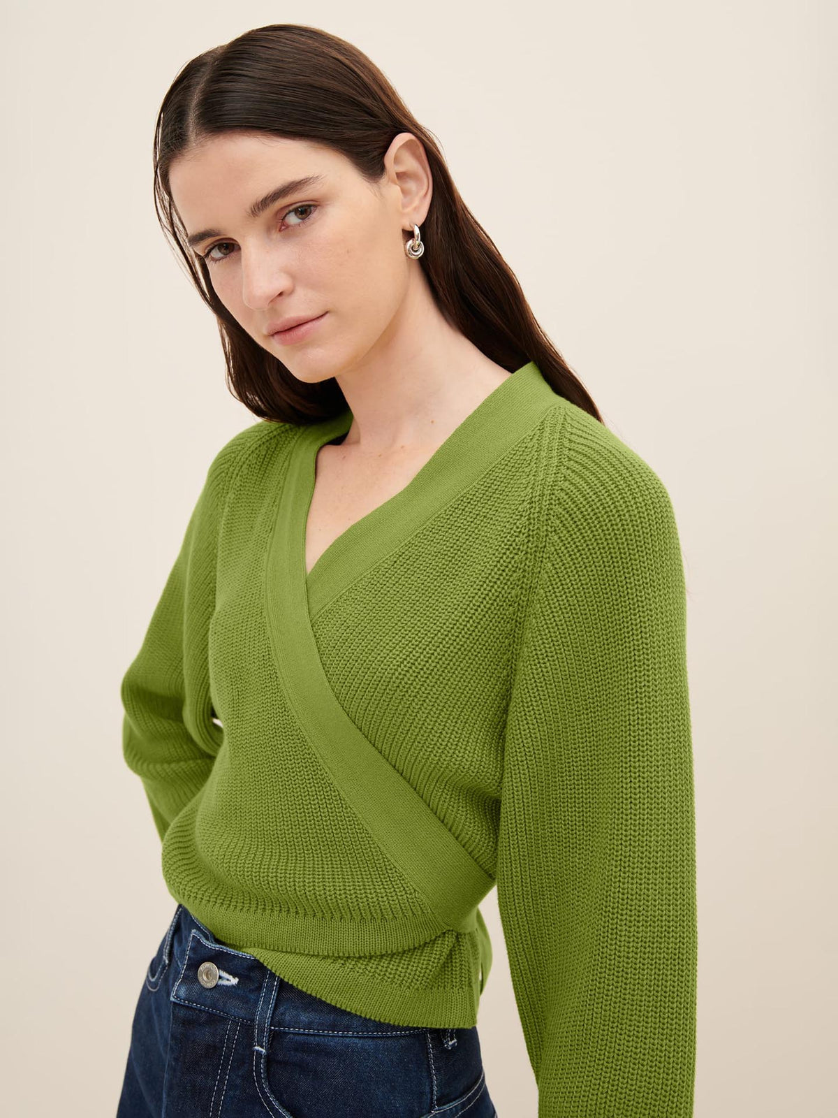 A person with long dark hair is wearing a Kowtow Composure Cardigan in Leaf, paired with dark jeans, standing against a plain beige background.