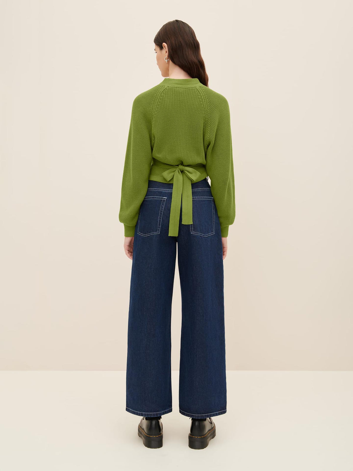 Person standing with their back to the camera, wearing a Kowtow Composure Cardigan in Leaf and wide-leg denim pants.